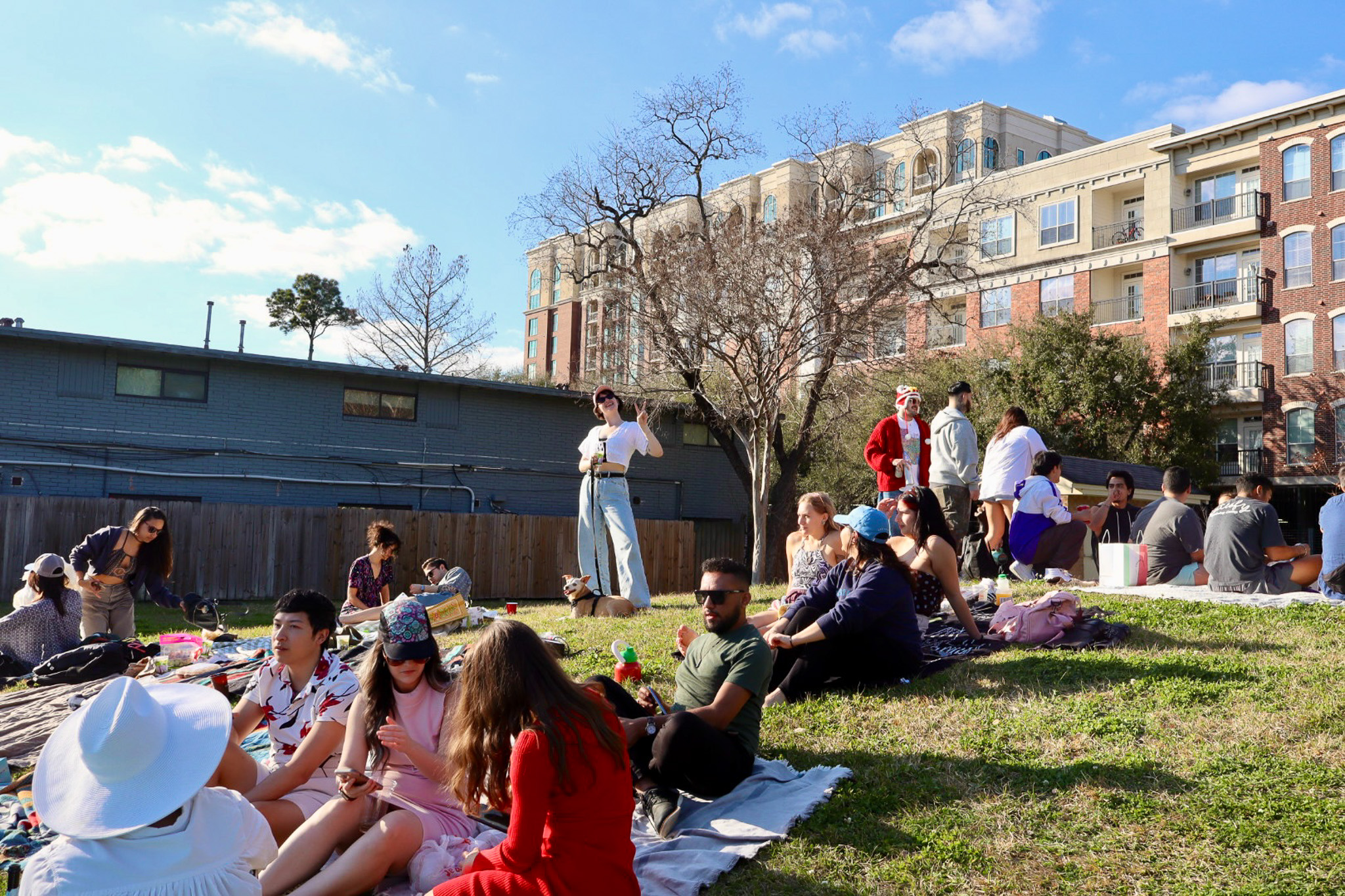 Group of people relaxing and socializing on blankets in a sunny grassy area with urban buildings in the background.