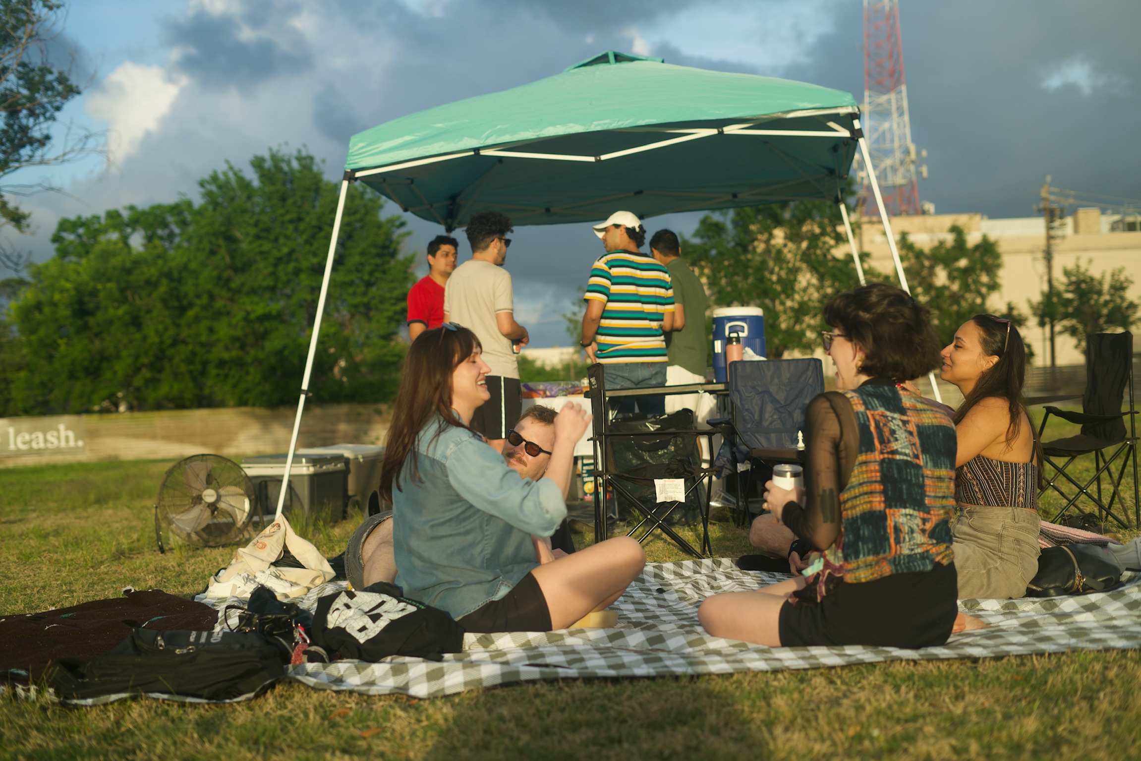People enjoying a sunny picnic on blankets with drinks and snacks in a park, with apartment buildings in the background.