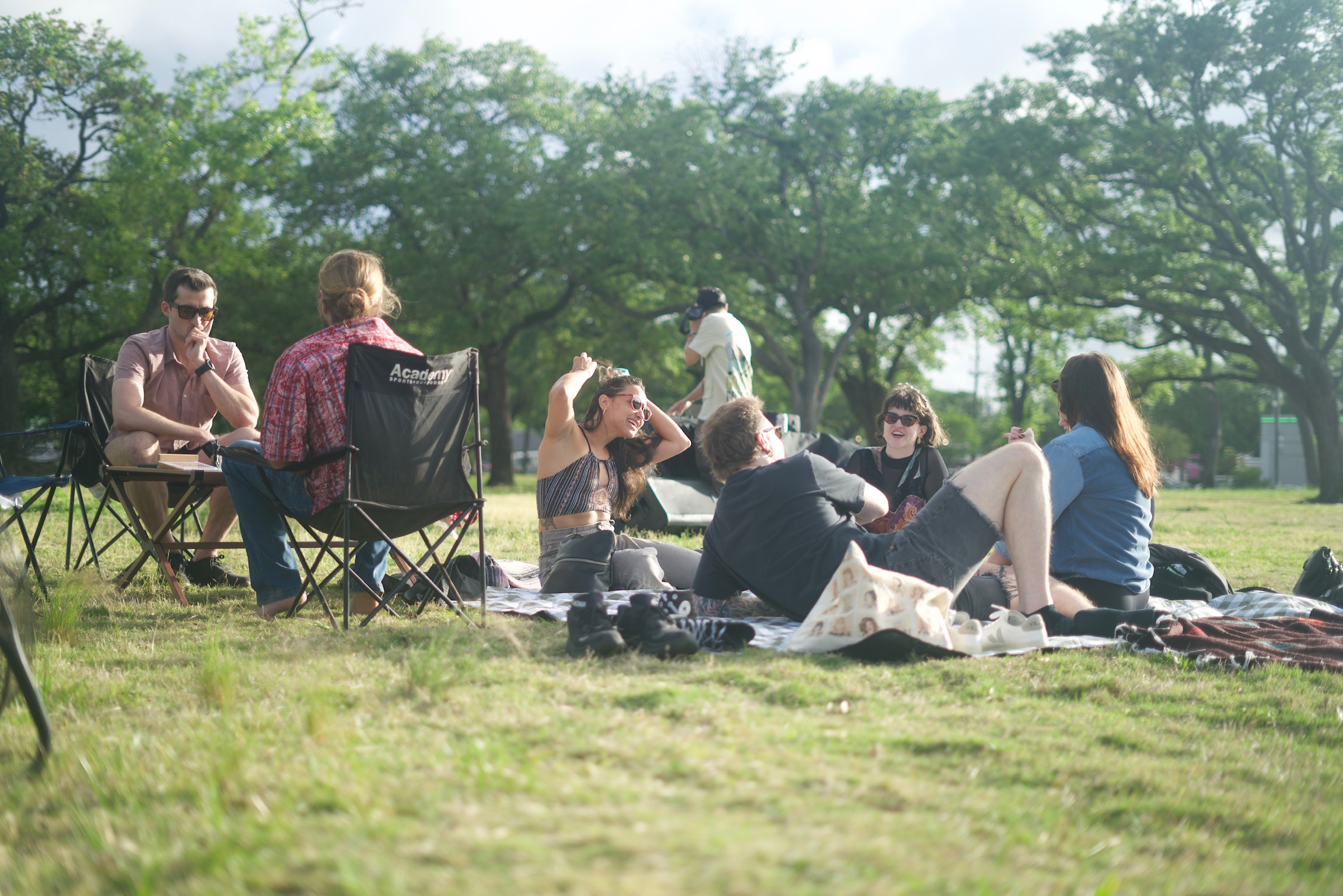Group of people relaxing and socializing on blankets in a sunny grassy area with urban buildings in the background.