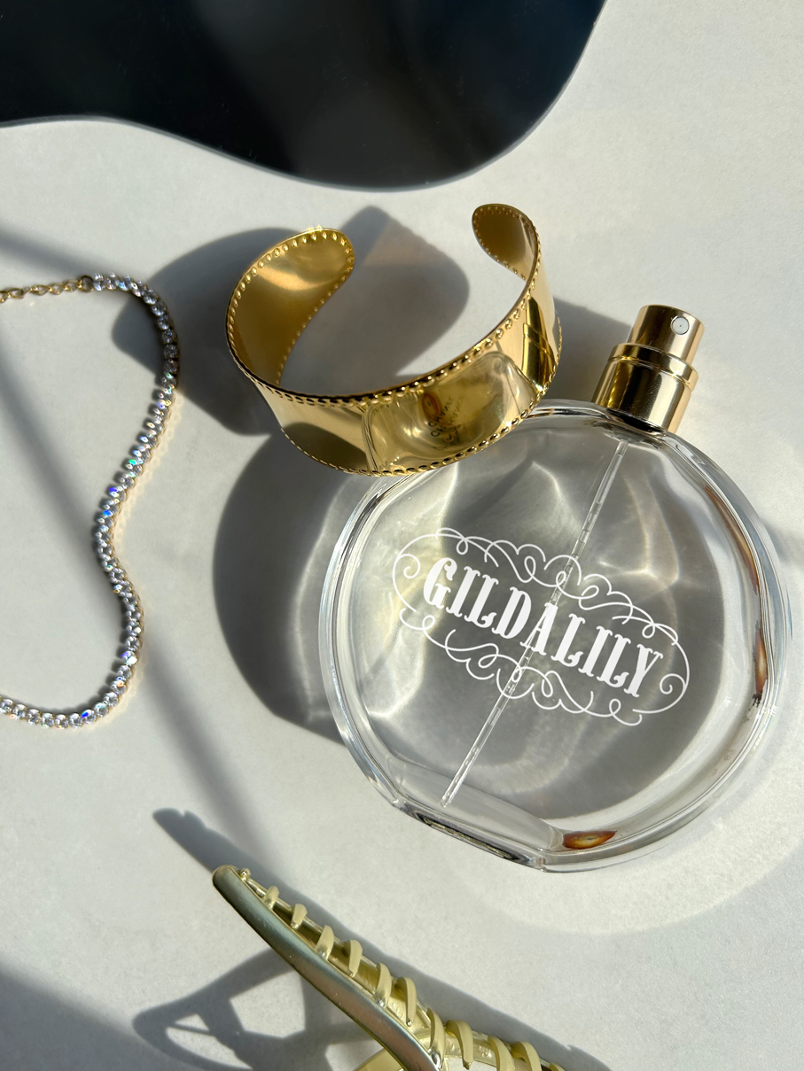 Clear Gildalily perfume bottle, gold cuff bracelet, rhinestone chain, and gold hair clip on white surface.
