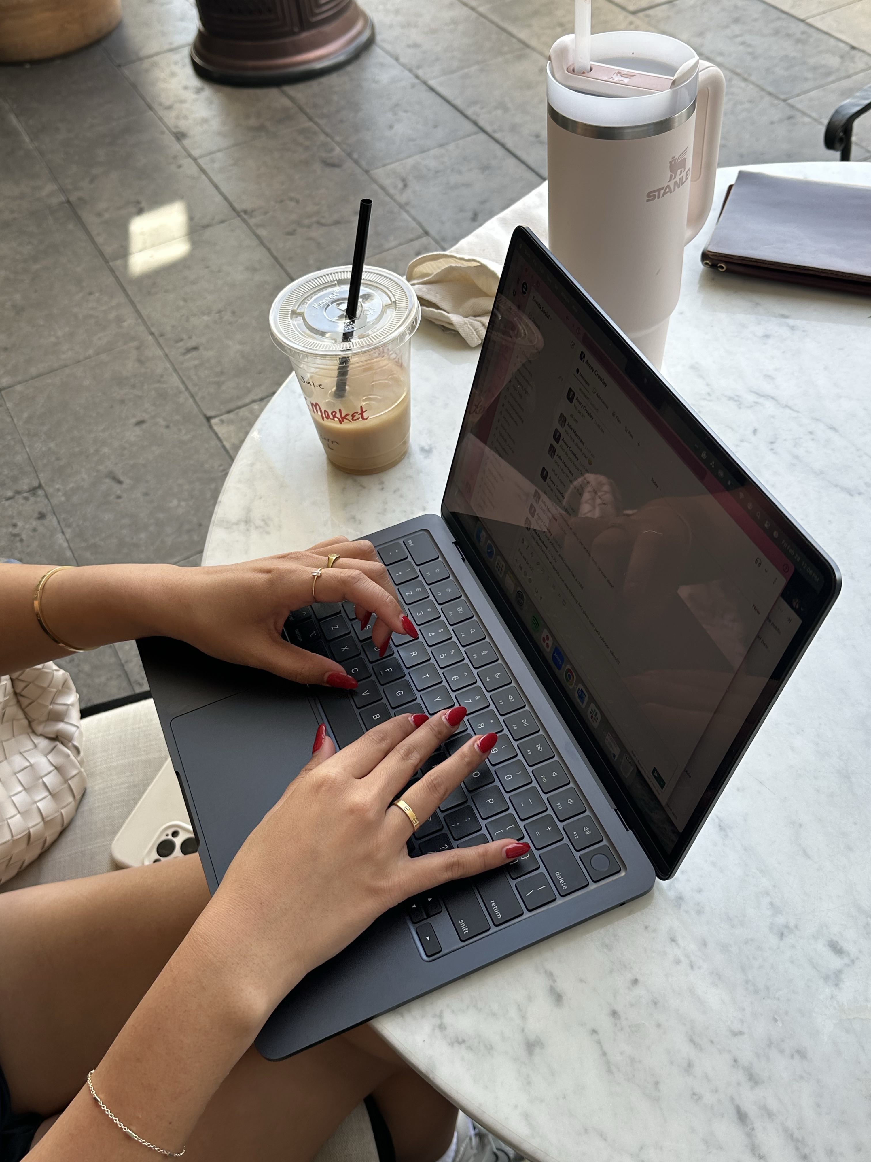 Person with red nail polish typing on a laptop at a marble table with drinks nearby.