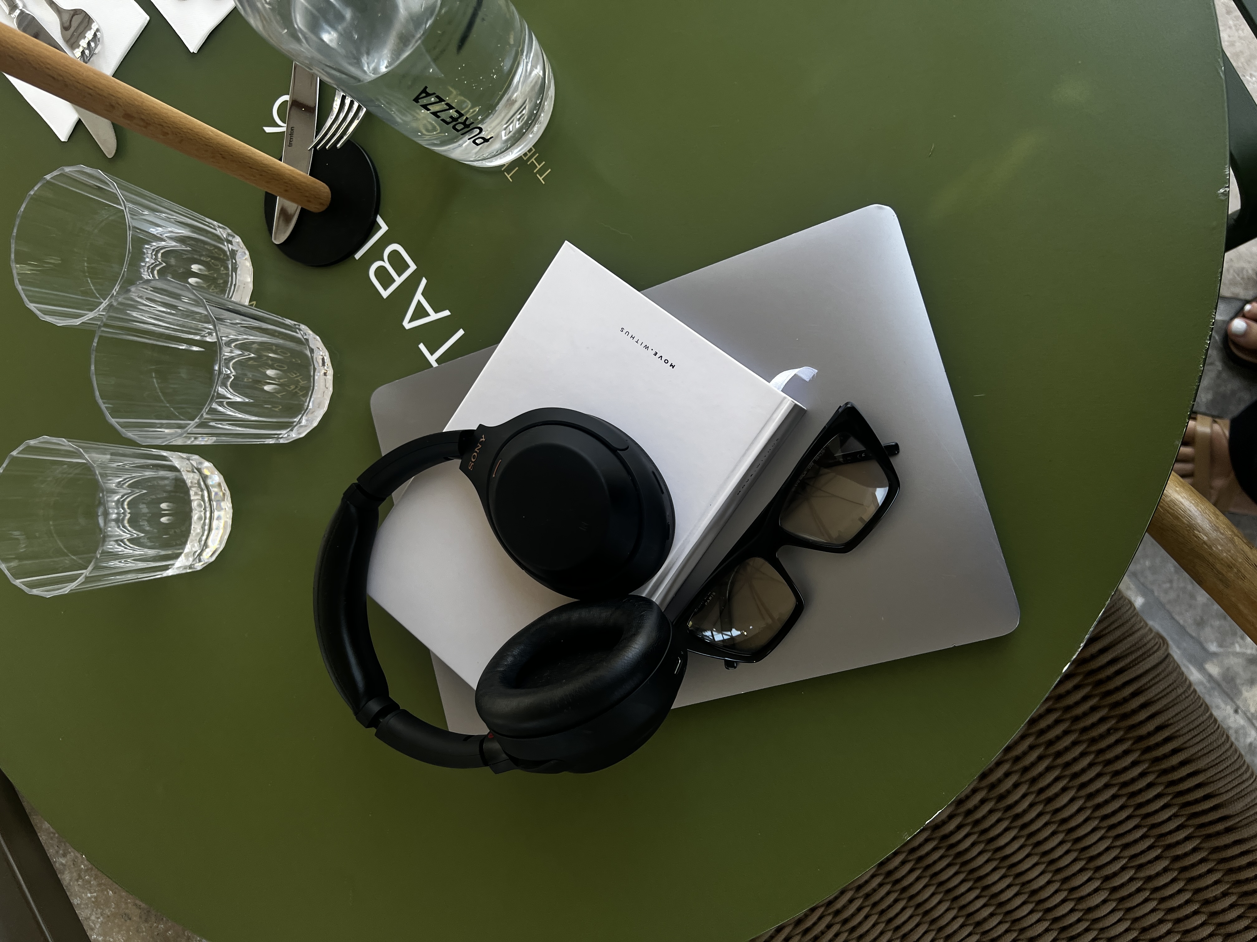 Black headphones, sunglasses, and a white book on closed silver laptop on green table.