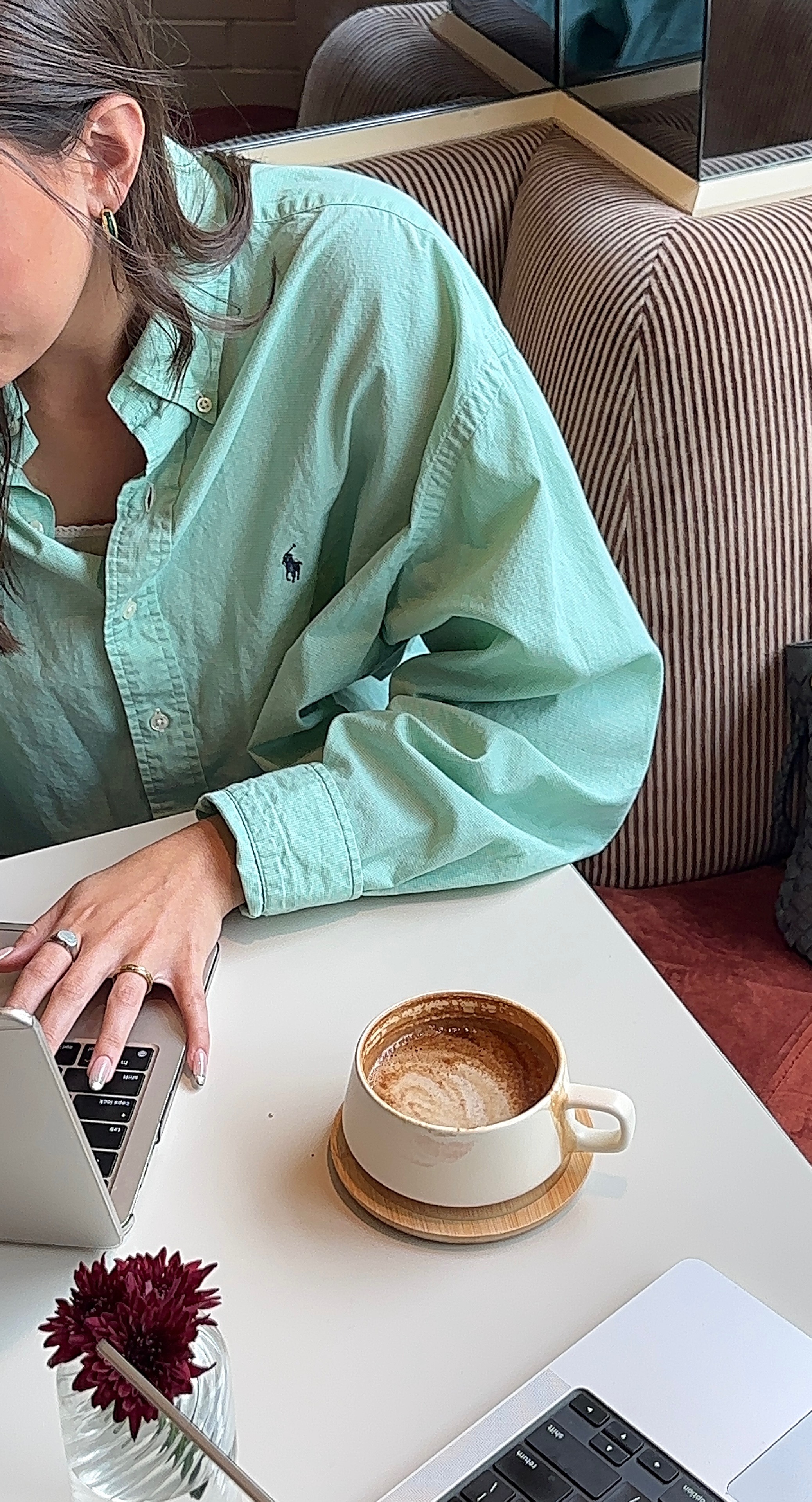Person in mint green shirt using laptop next to coffee cup and flower on white table.