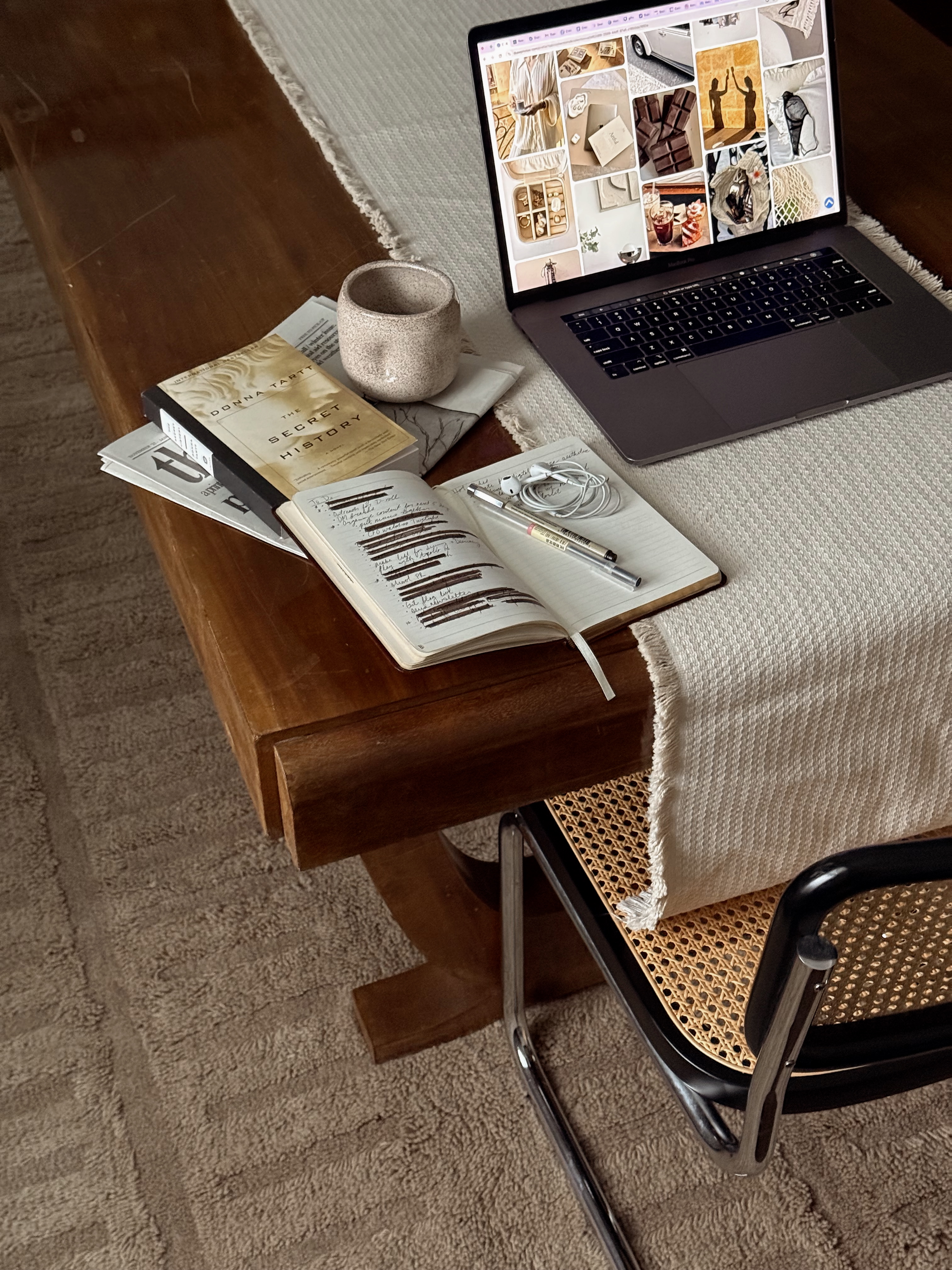 Wooden table with a laptop, notebook with pen and earphones, book, mug, and newspapers on a beige cloth.