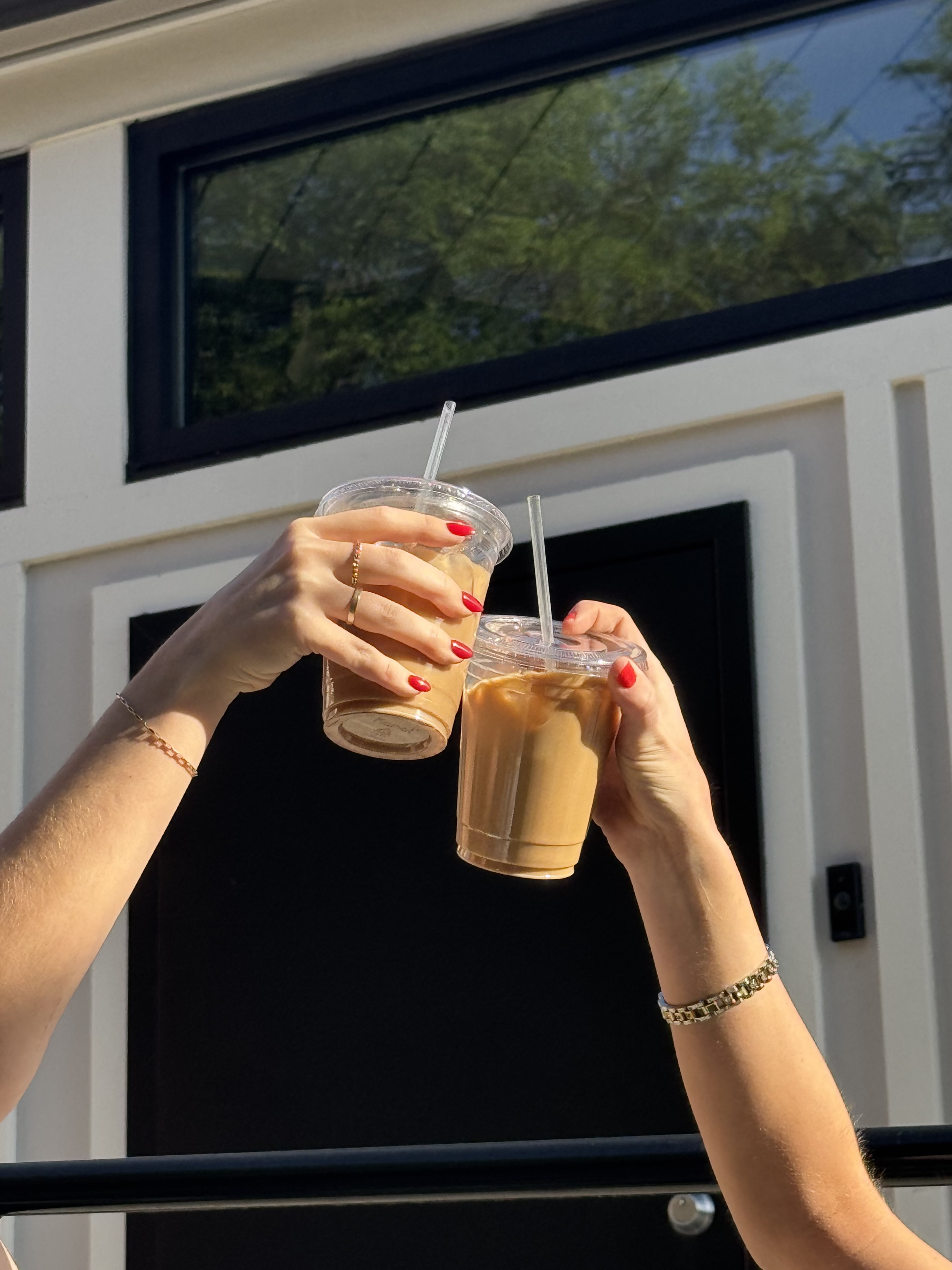 Two hands with red nails holding iced coffee cups with straws raised in a toast outdoors.