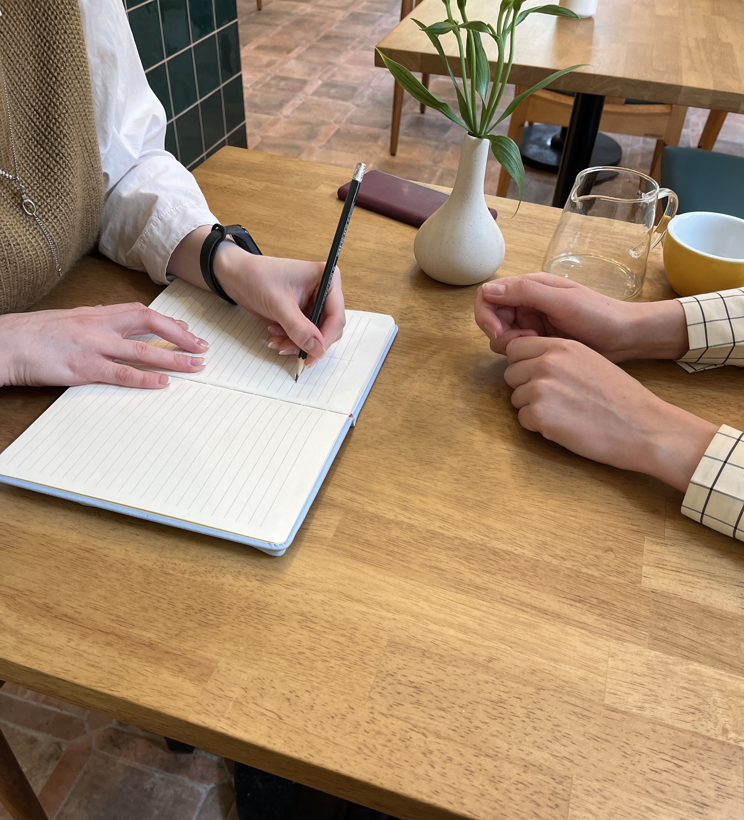 Two people at a wooden table, one writing in a notebook, the other resting hands nearby.