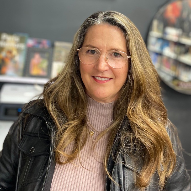 Smiling woman with long wavy hair wearing glasses, a pink turtleneck, and a black leather jacket.