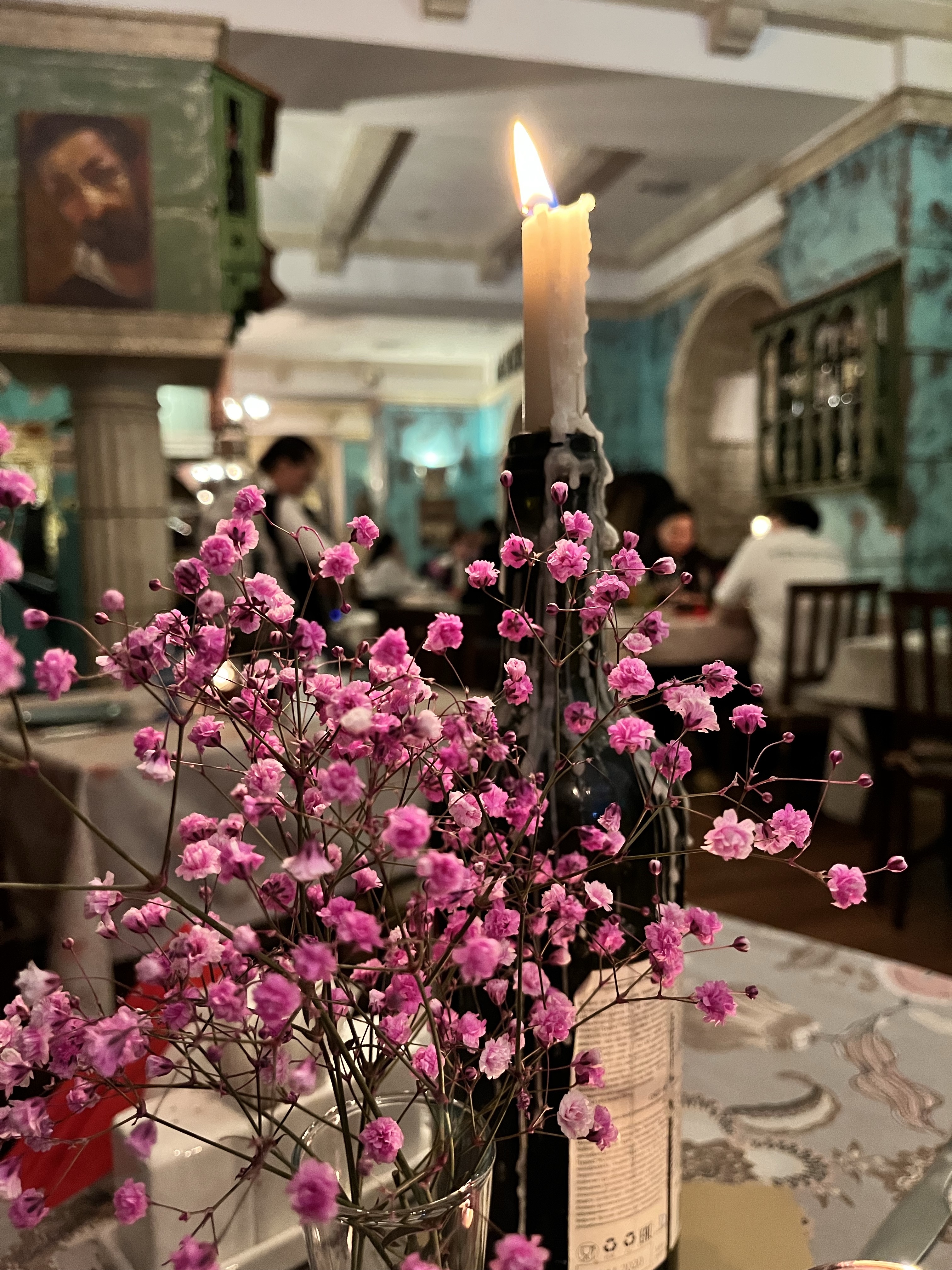 Pink flowers in a glass next to a lit candle on a restaurant table with blurred diners and blue walls.