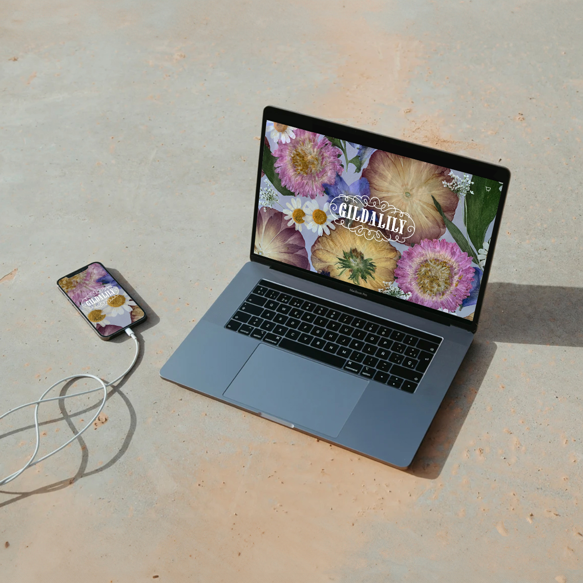 Open laptop and smartphone on concrete floor showing Gildalily floral design on screens.