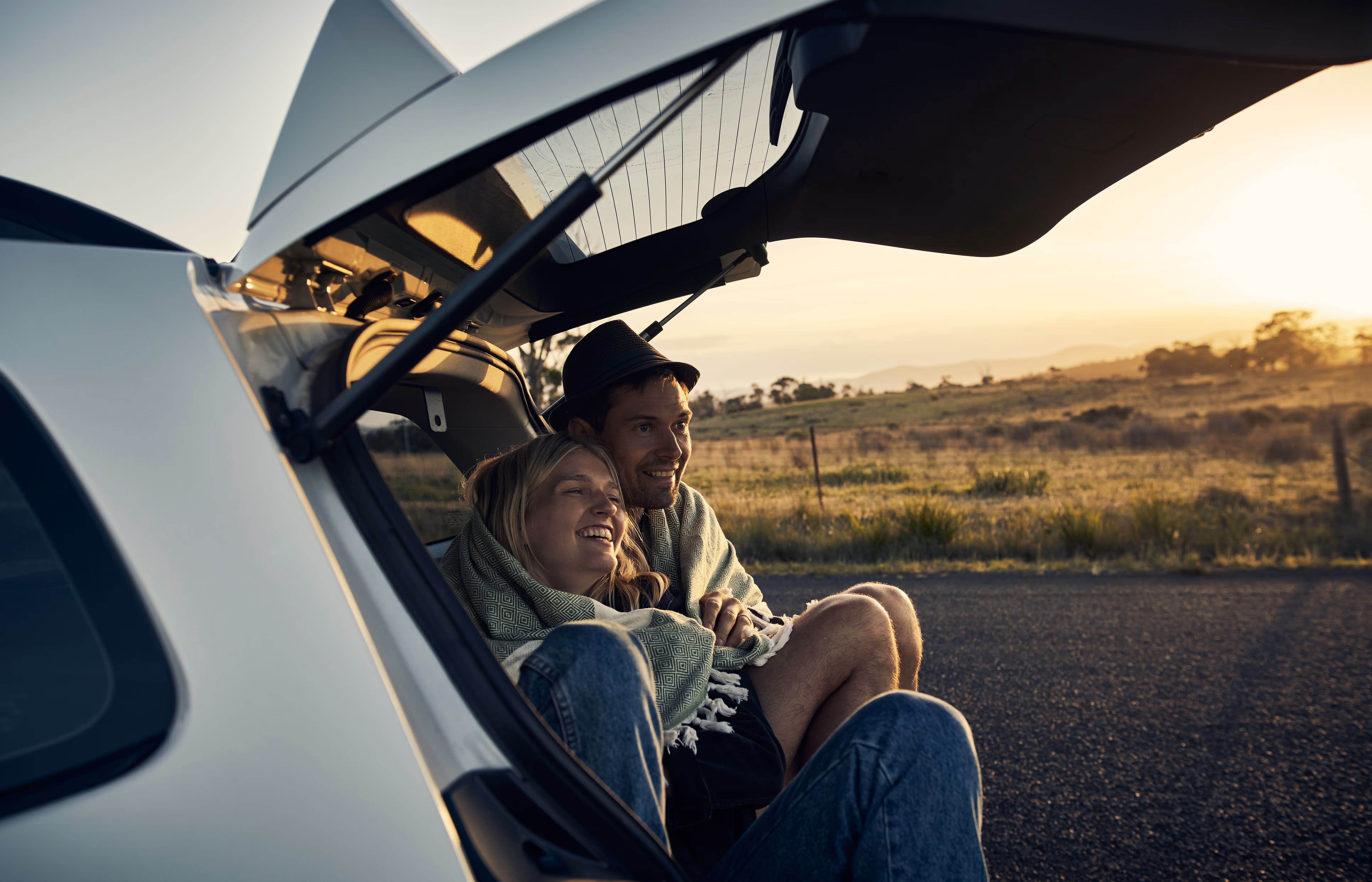 Couple wrapped in a blanket sitting in the open rear of a car at sunset on a rural road.