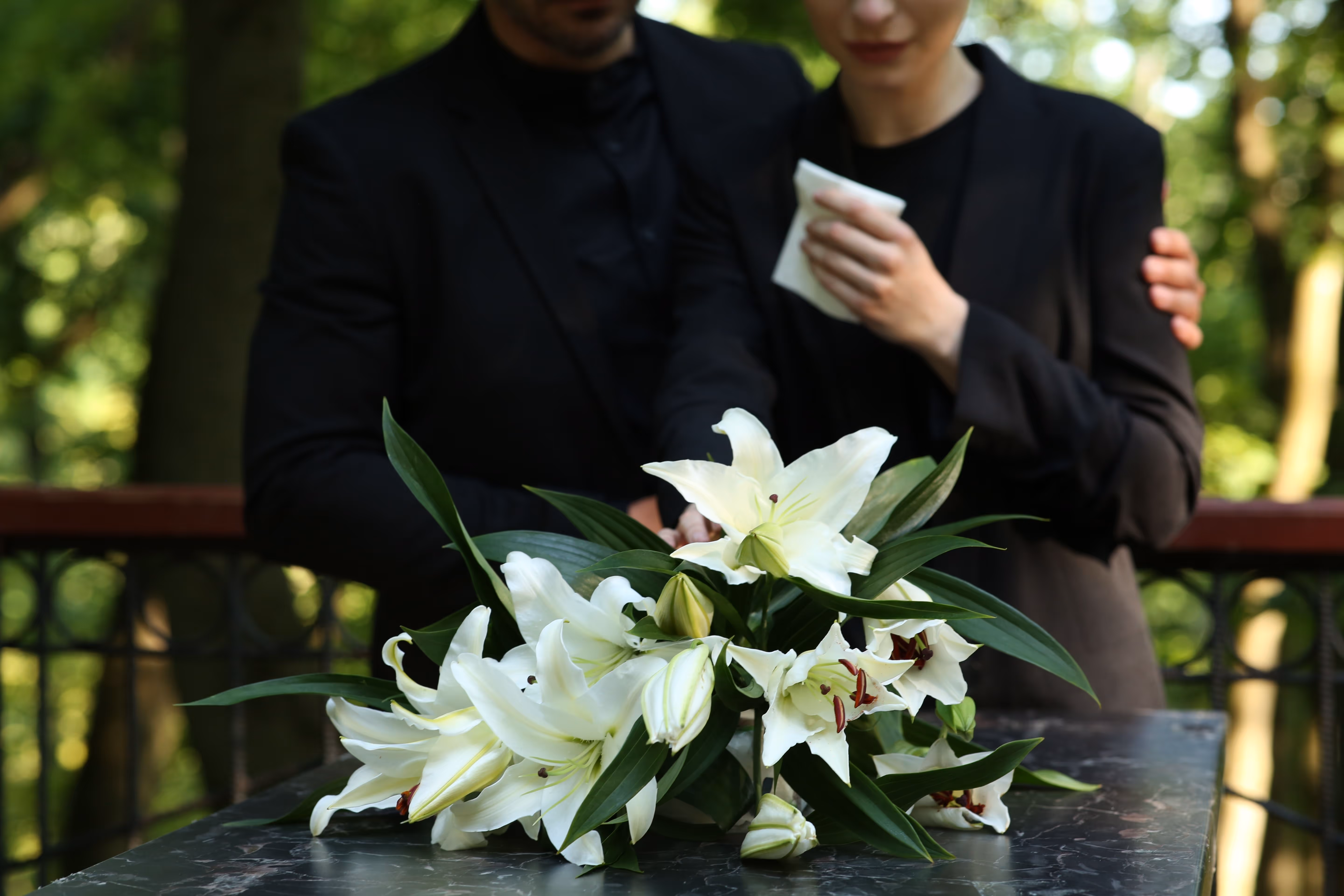White lilies on a marble table with two people dressed in black mourning in the background.