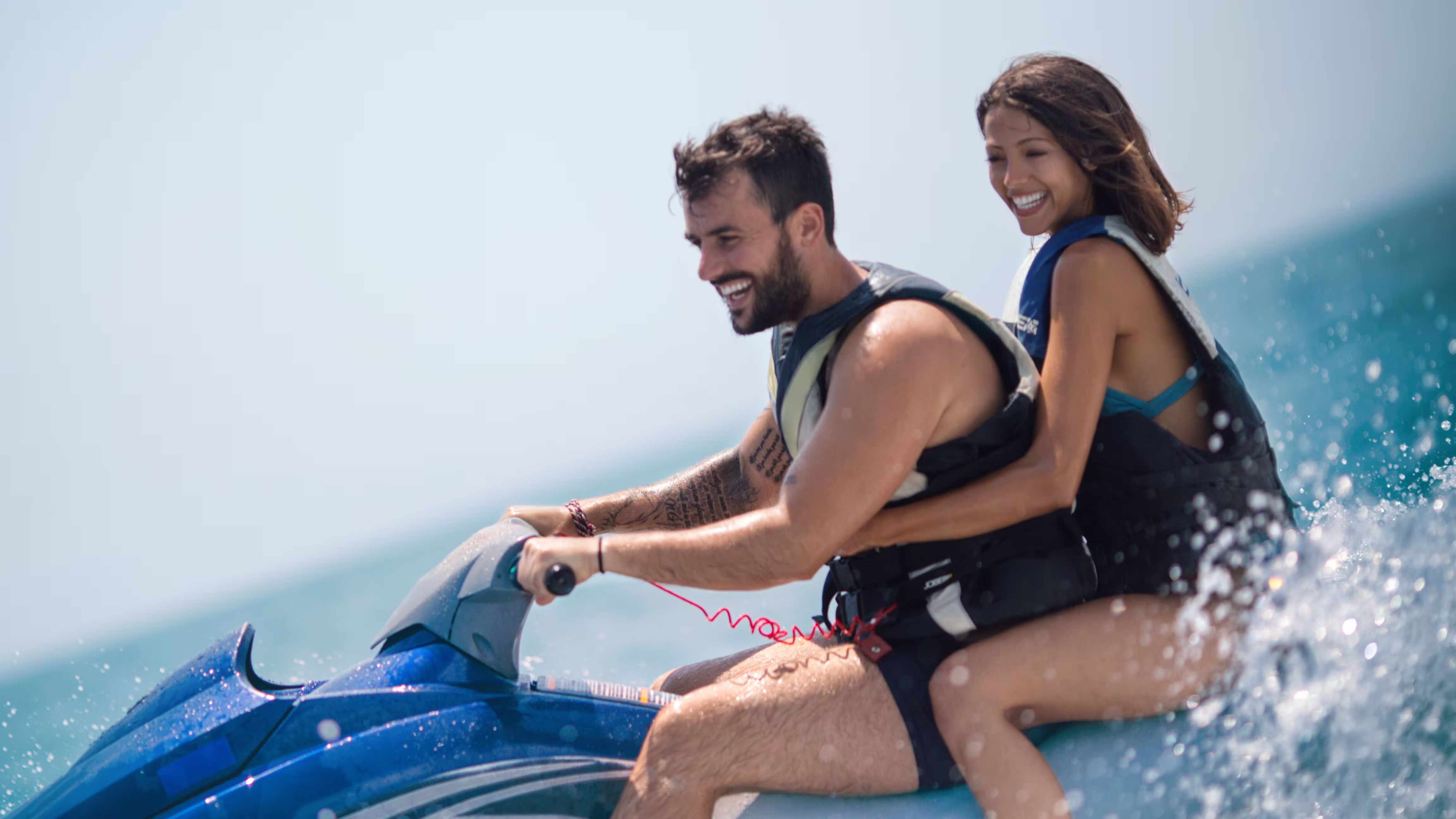 Smiling couple wearing life jackets riding a blue jet ski on the ocean with water splashing around.