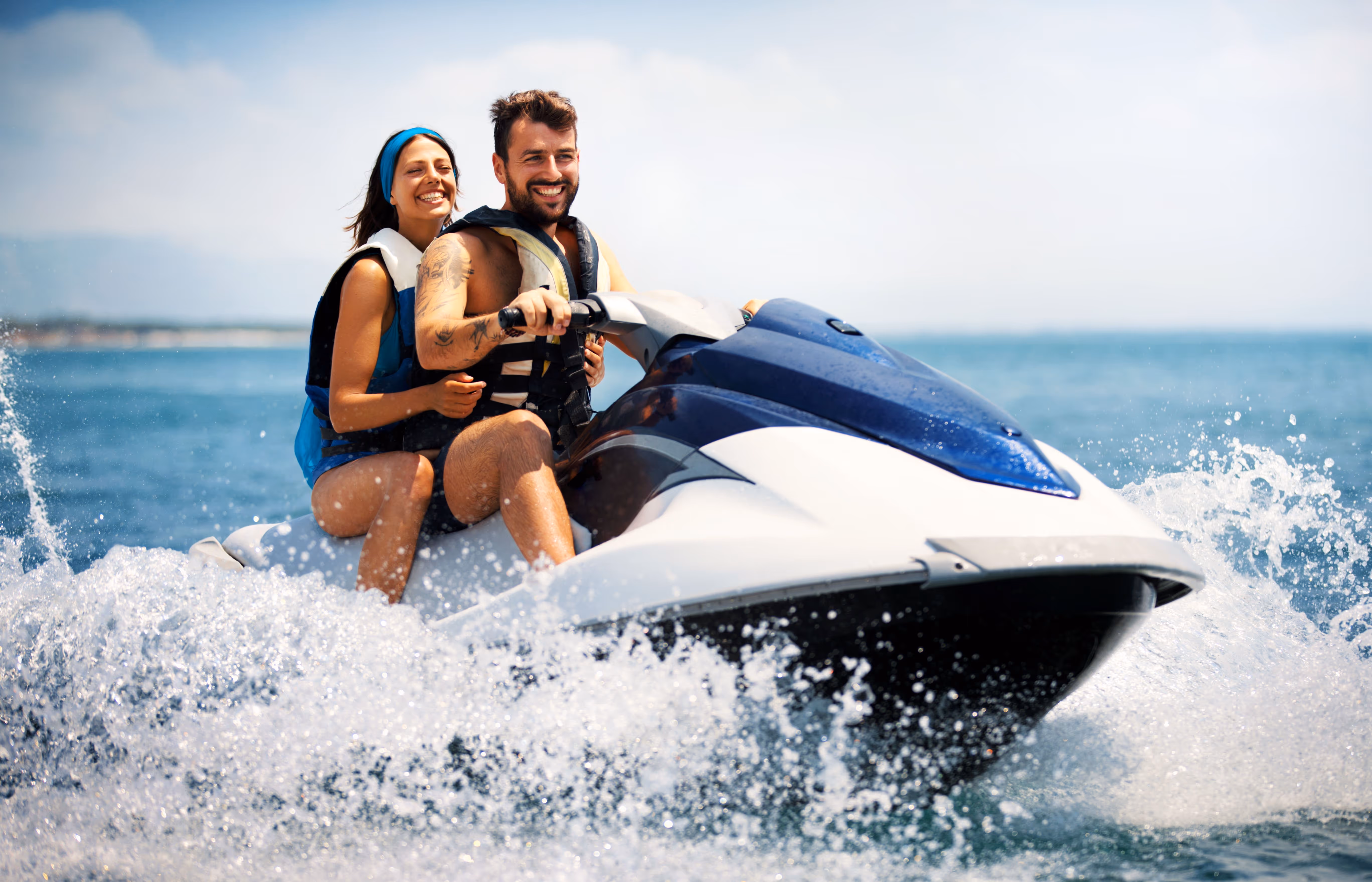 Smiling couple wearing life jackets riding a blue and white jet ski on the ocean with water splashing around.