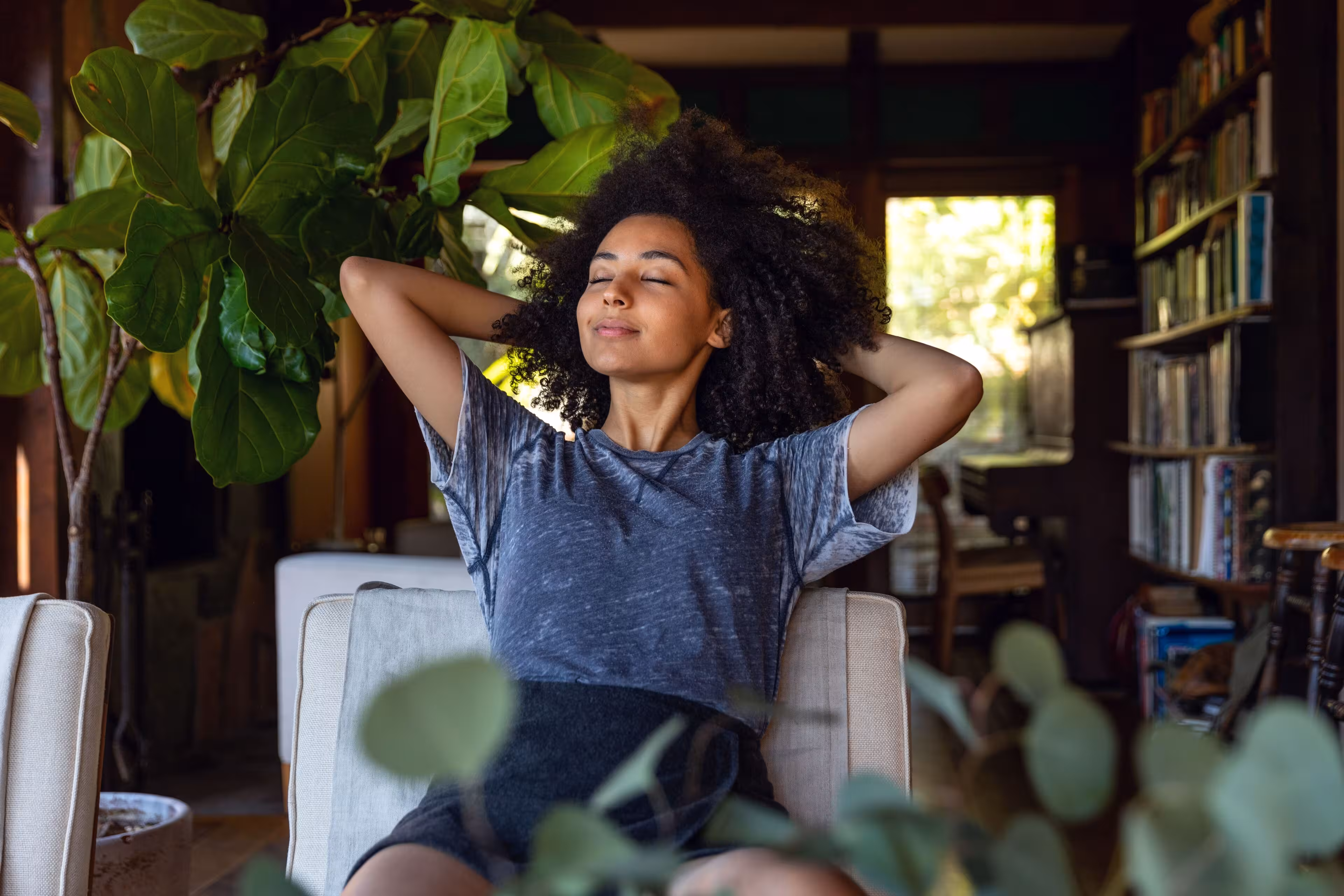 Young woman with curly hair sitting relaxed on a couch in a cozy room with plants and bookshelves.