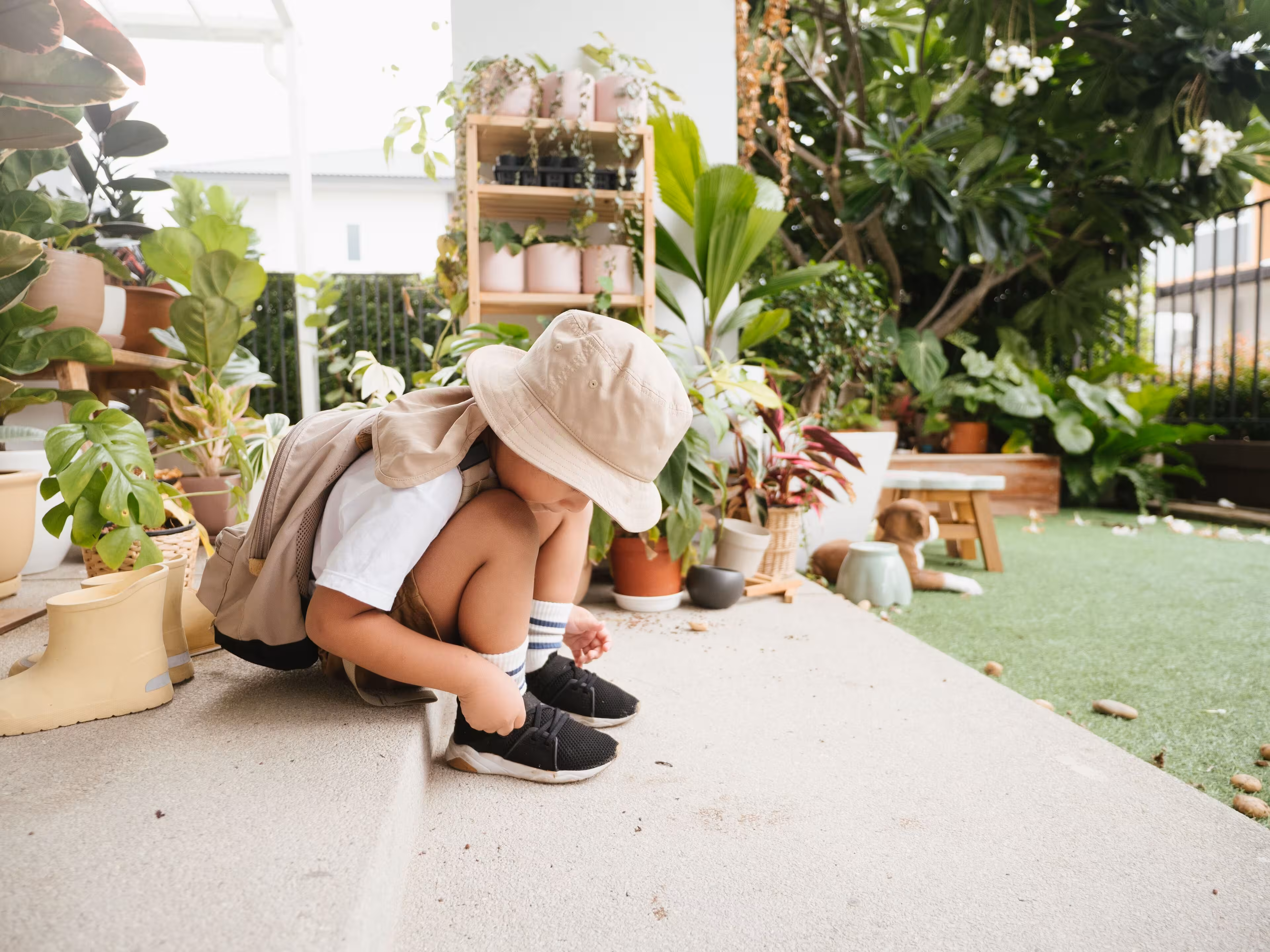 Child wearing a beige hat and backpack tying black shoes while sitting on outdoor steps surrounded by plants.