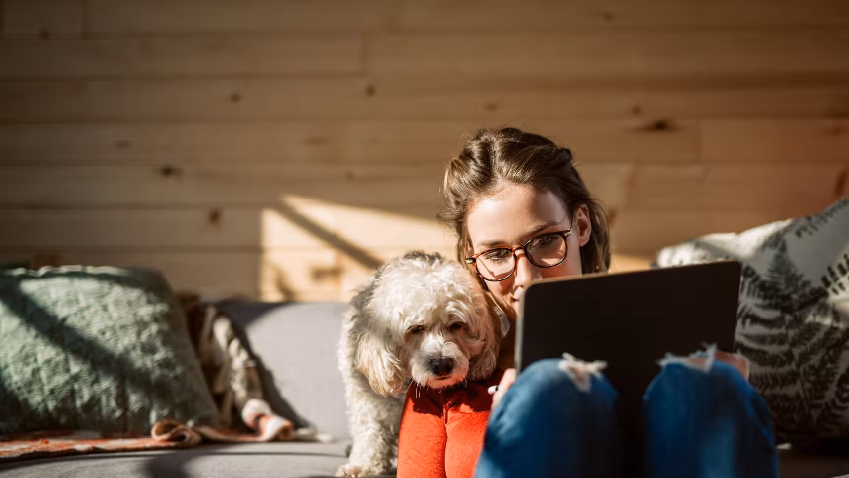 Woman with glasses sitting on a couch using a tablet while a fluffy dog rests its head on her knees.