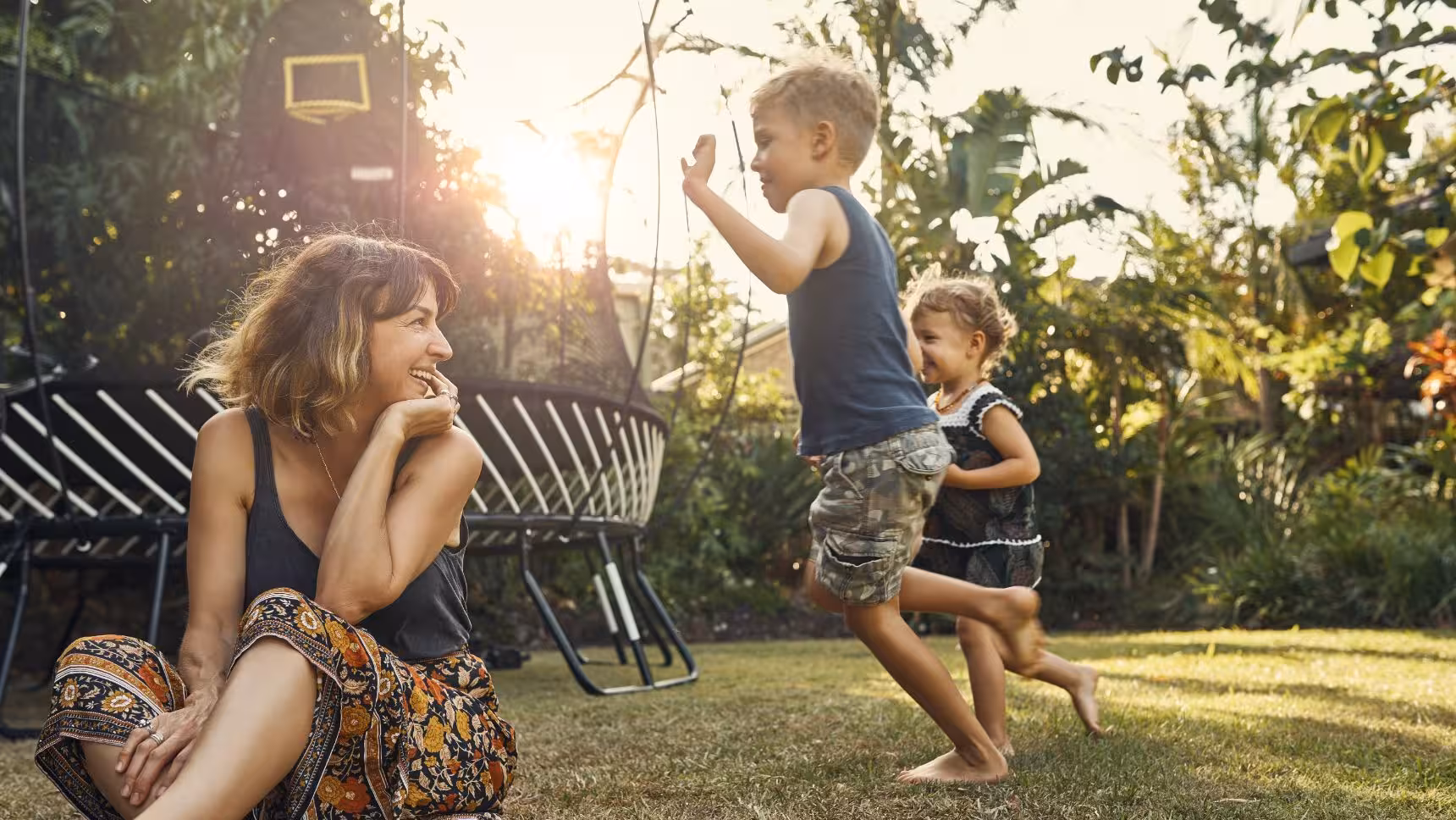 Woman sitting on grass smiling at two children playing and running barefoot in a sunny backyard.