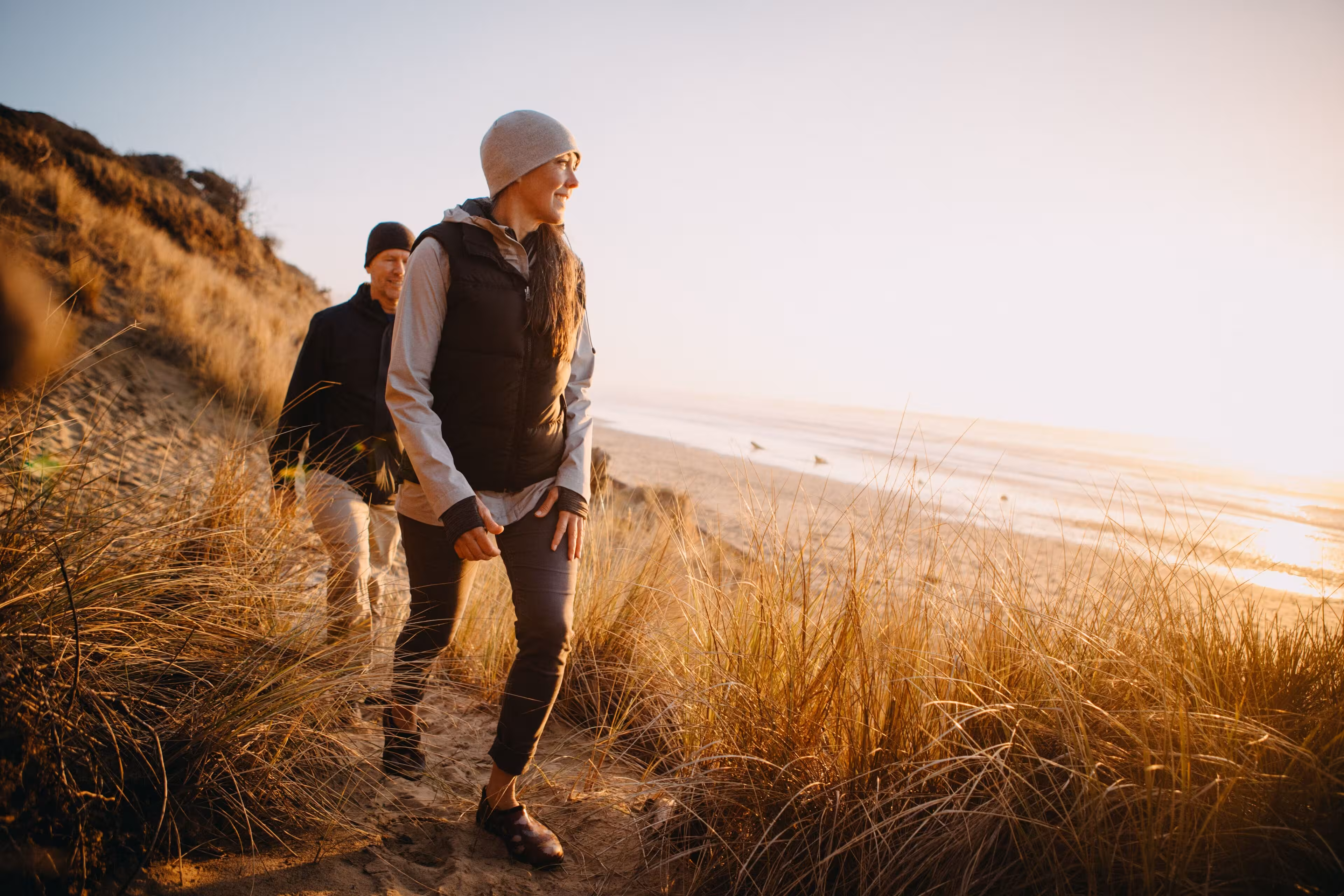 Couple hiking along a sandy beach path with tall grasses during a golden sunset.