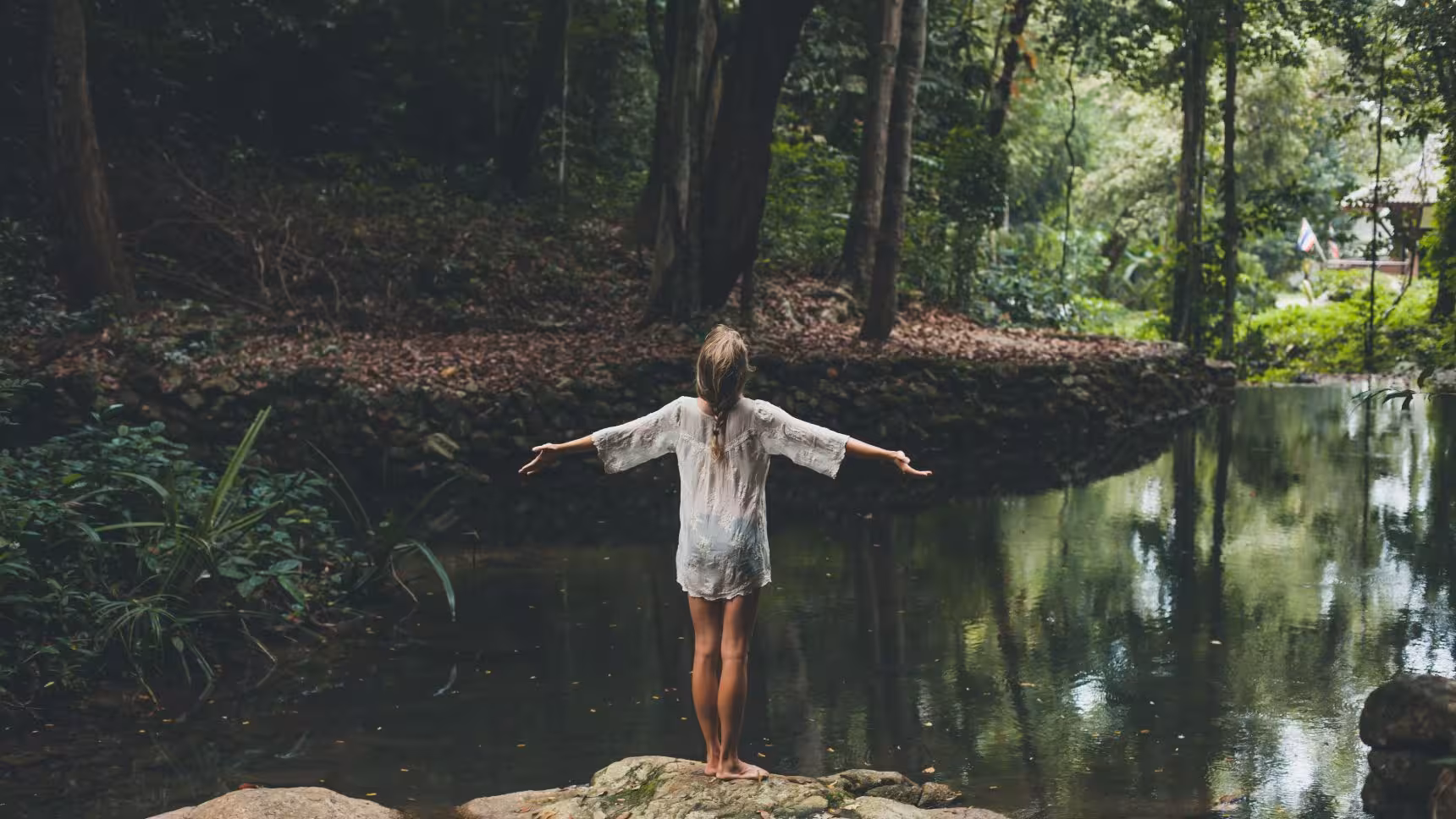 Woman standing barefoot on a rock with arms outstretched in front of a calm creek surrounded by dense forest.