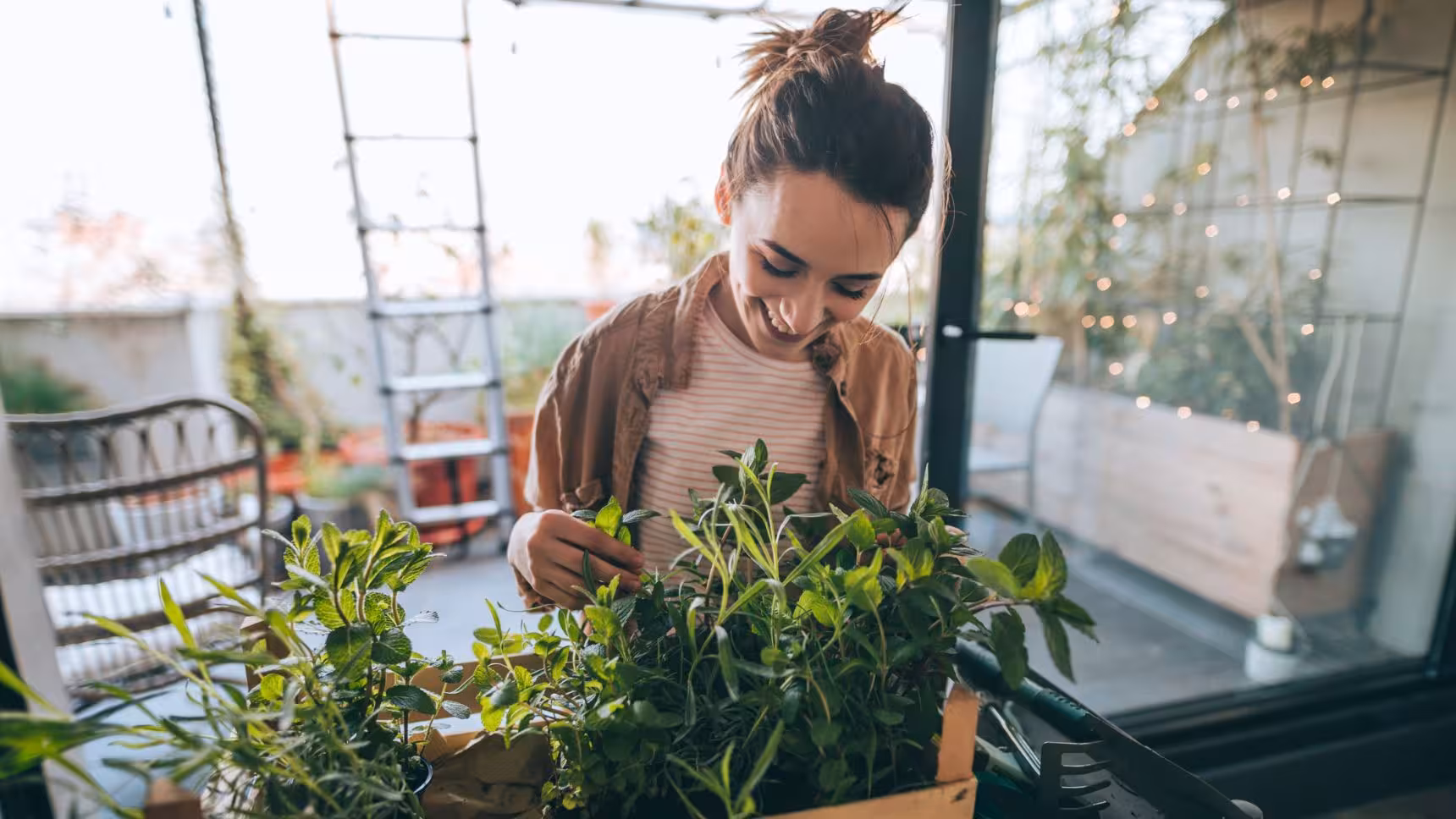 Smiling woman tending to green plants on a balcony garden with gardening tools nearby.