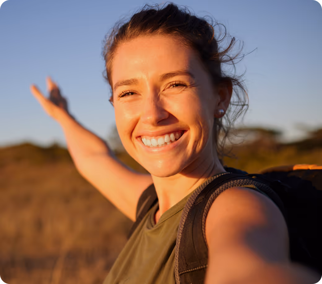 Smiling woman with a backpack extending her arm outdoors at sunset.