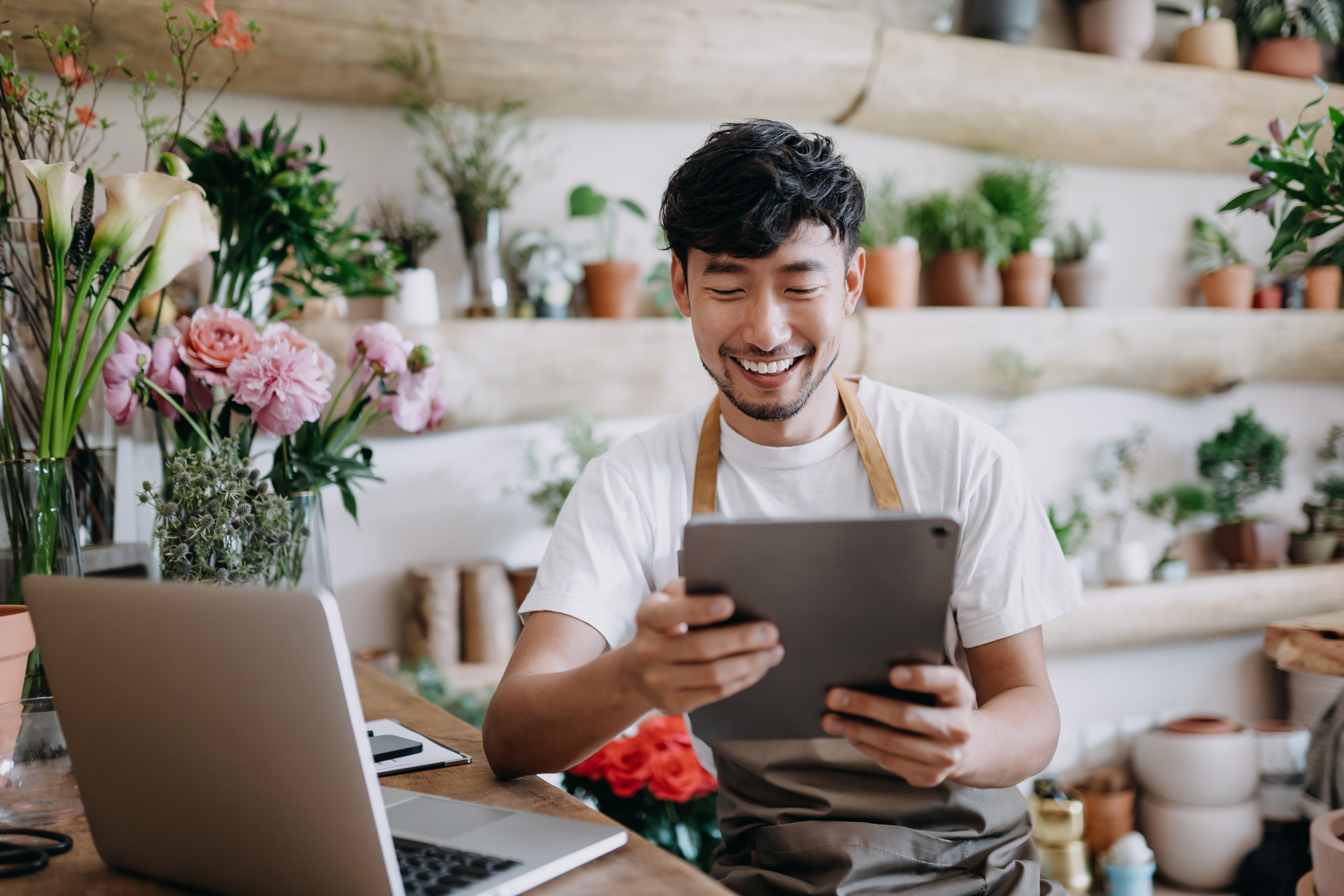 Smiling man in an apron using a tablet in a flower shop with plants and flowers around.