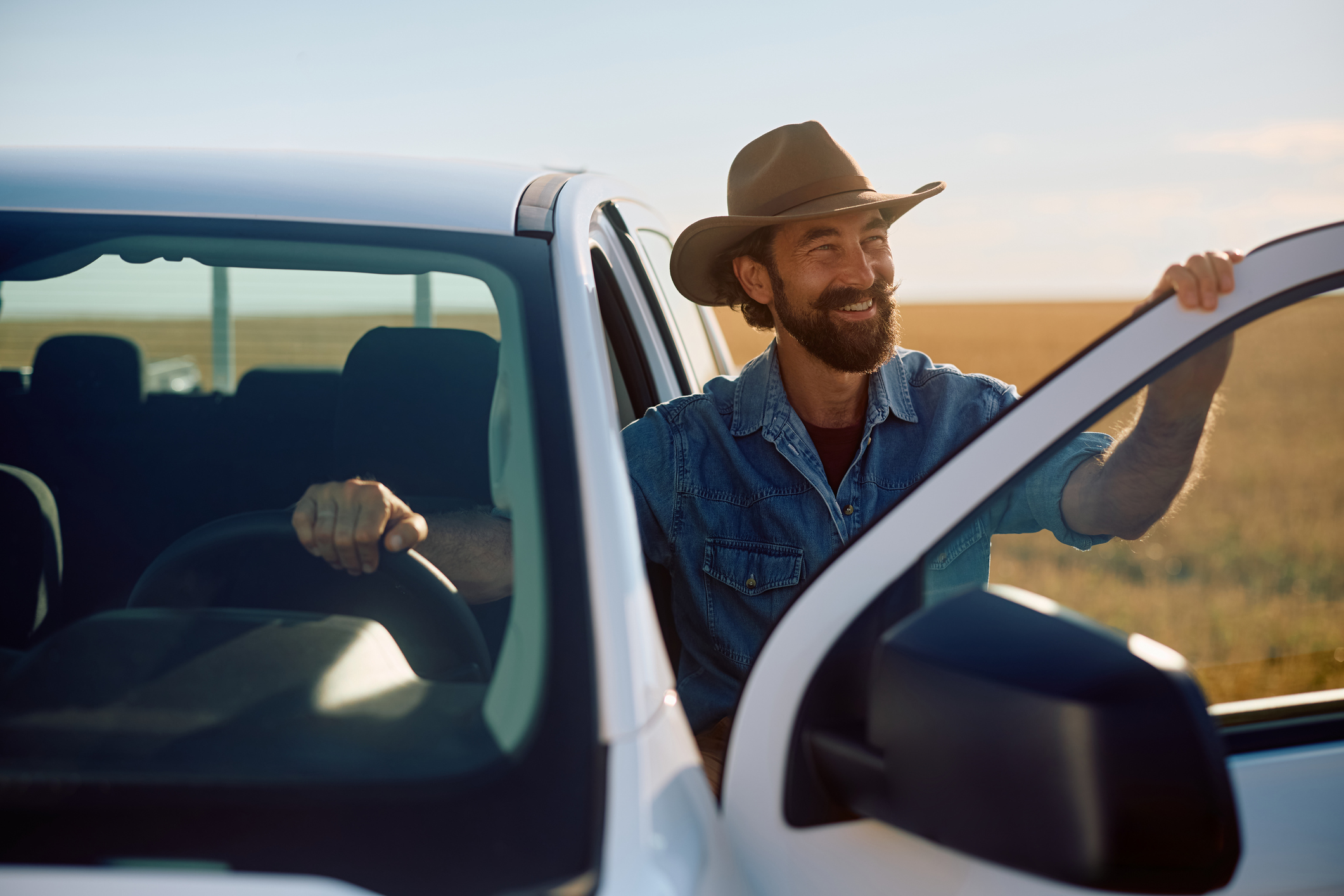 Smiling man wearing a cowboy hat and denim shirt opening the door of a white pickup truck on a farm.
