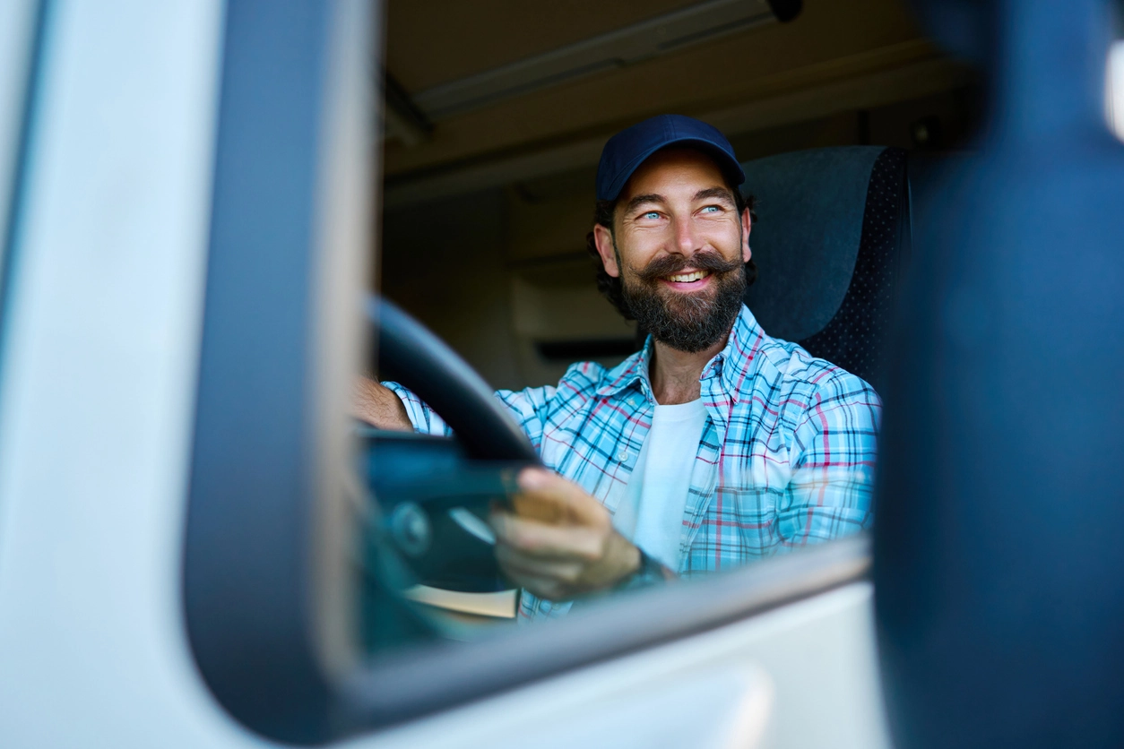 Smiling bearded man wearing a cap and plaid shirt driving a vehicle, seen through the side mirror.