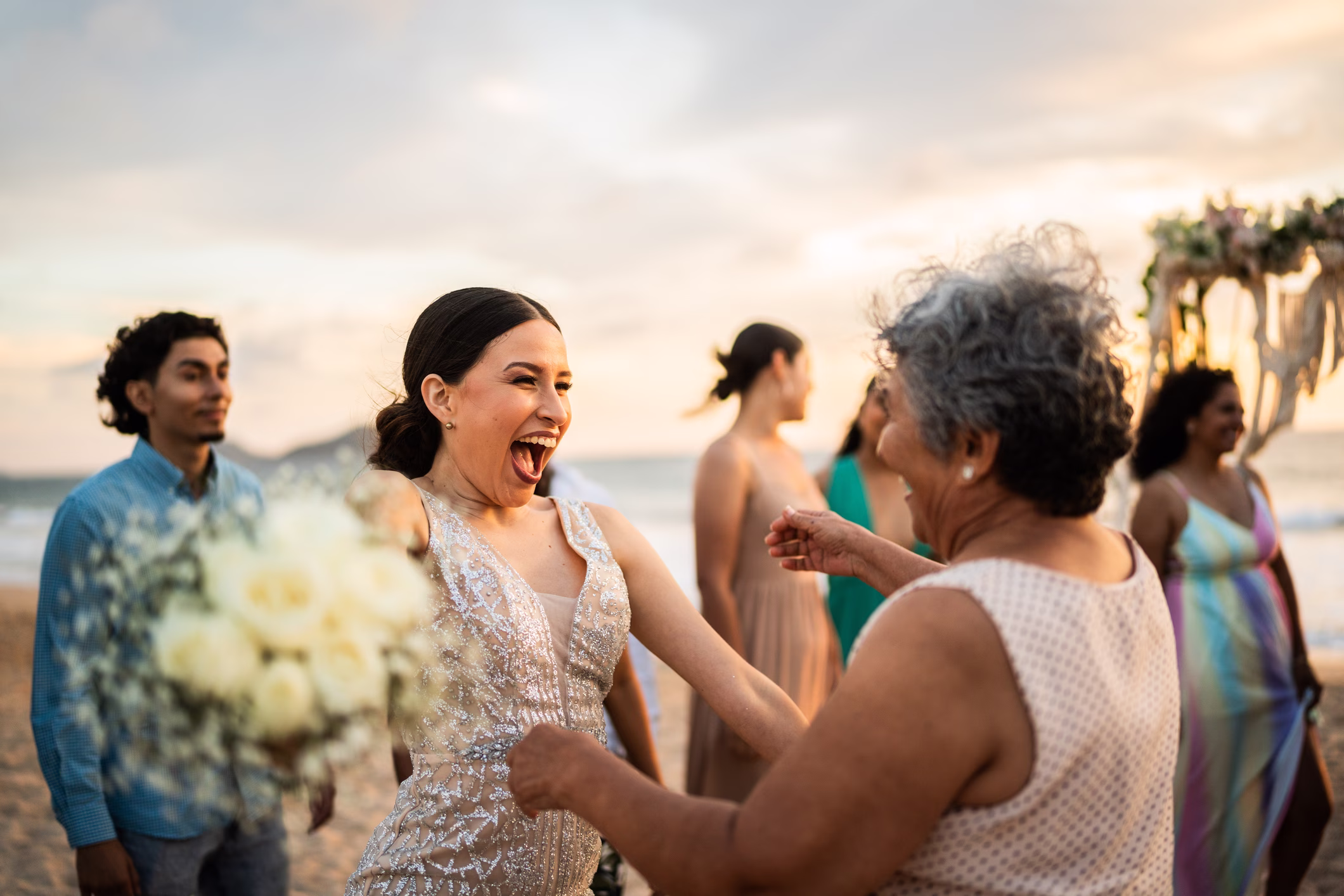 Bride in a beaded dress joyfully embraces an older woman on a beach with wedding guests in the background.
