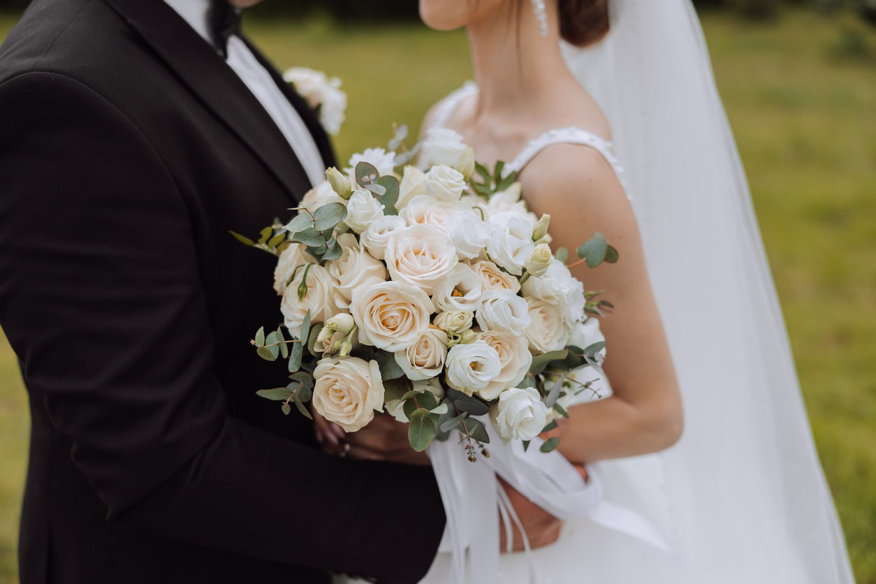 Bride and groom holding a bouquet of white and cream roses with eucalyptus leaves outdoors.