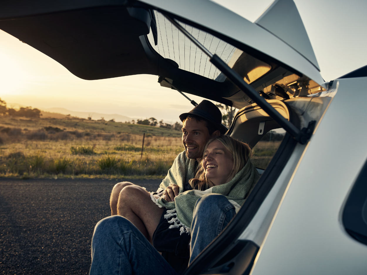 Happy couple wrapped in a blanket sitting together in a car trunk at sunset on a rural road.
