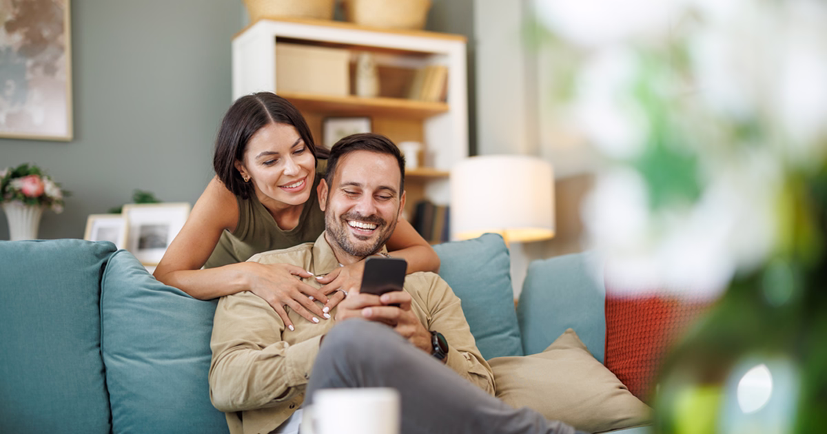 Smiling couple sitting on a couch, looking at a smartphone together in a cozy living room.