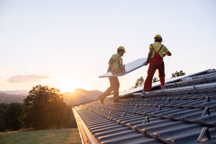 Two workers installing solar panels on a roof at sunset over a rural landscape.