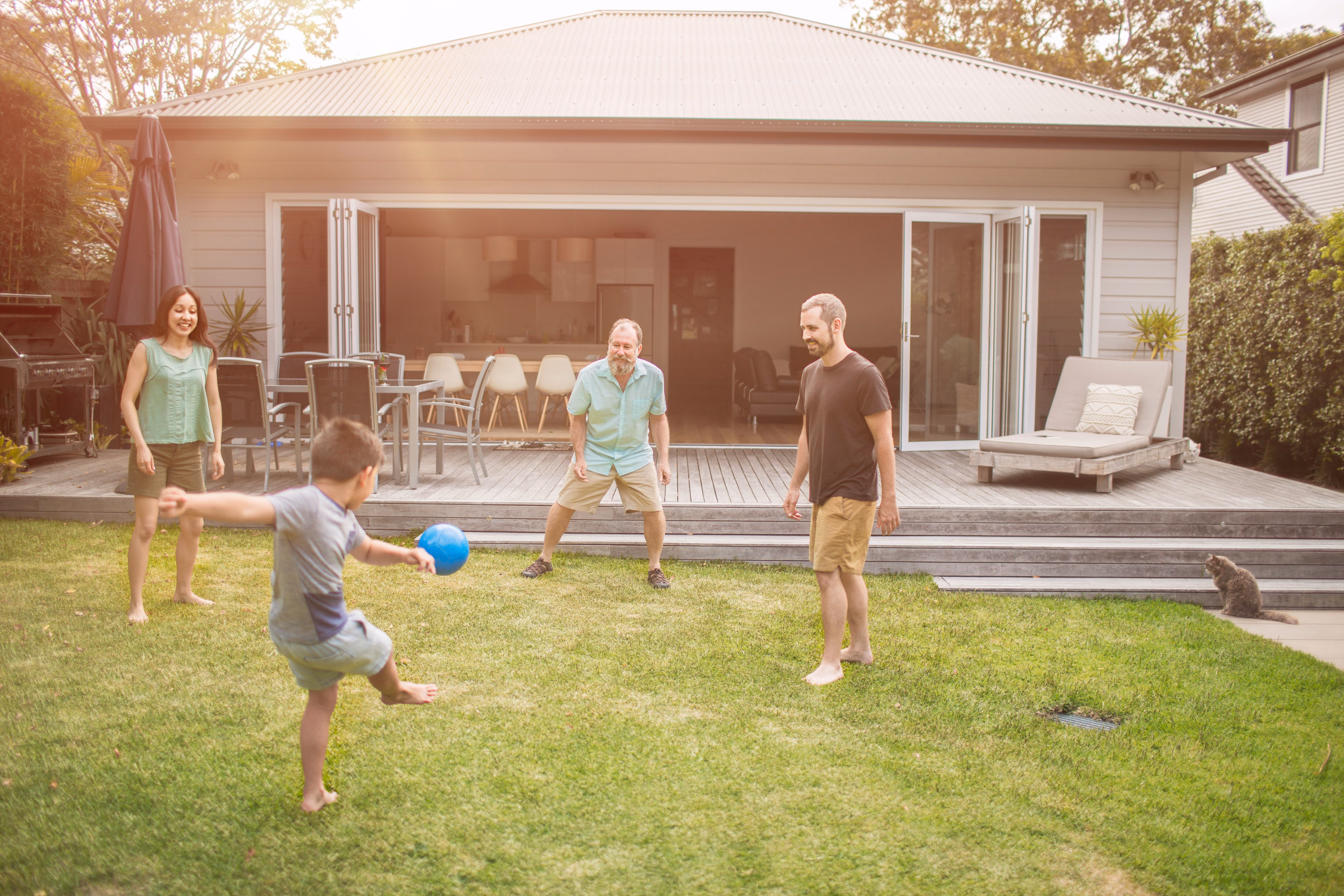 Young boy kicking a blue ball in the backyard with three adults watching and a cat on the side.