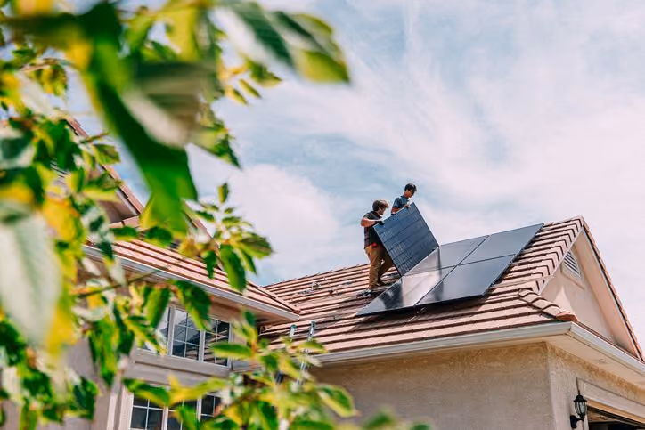 Two workers install solar panels on a house roof under a blue sky with some leaves in foreground.