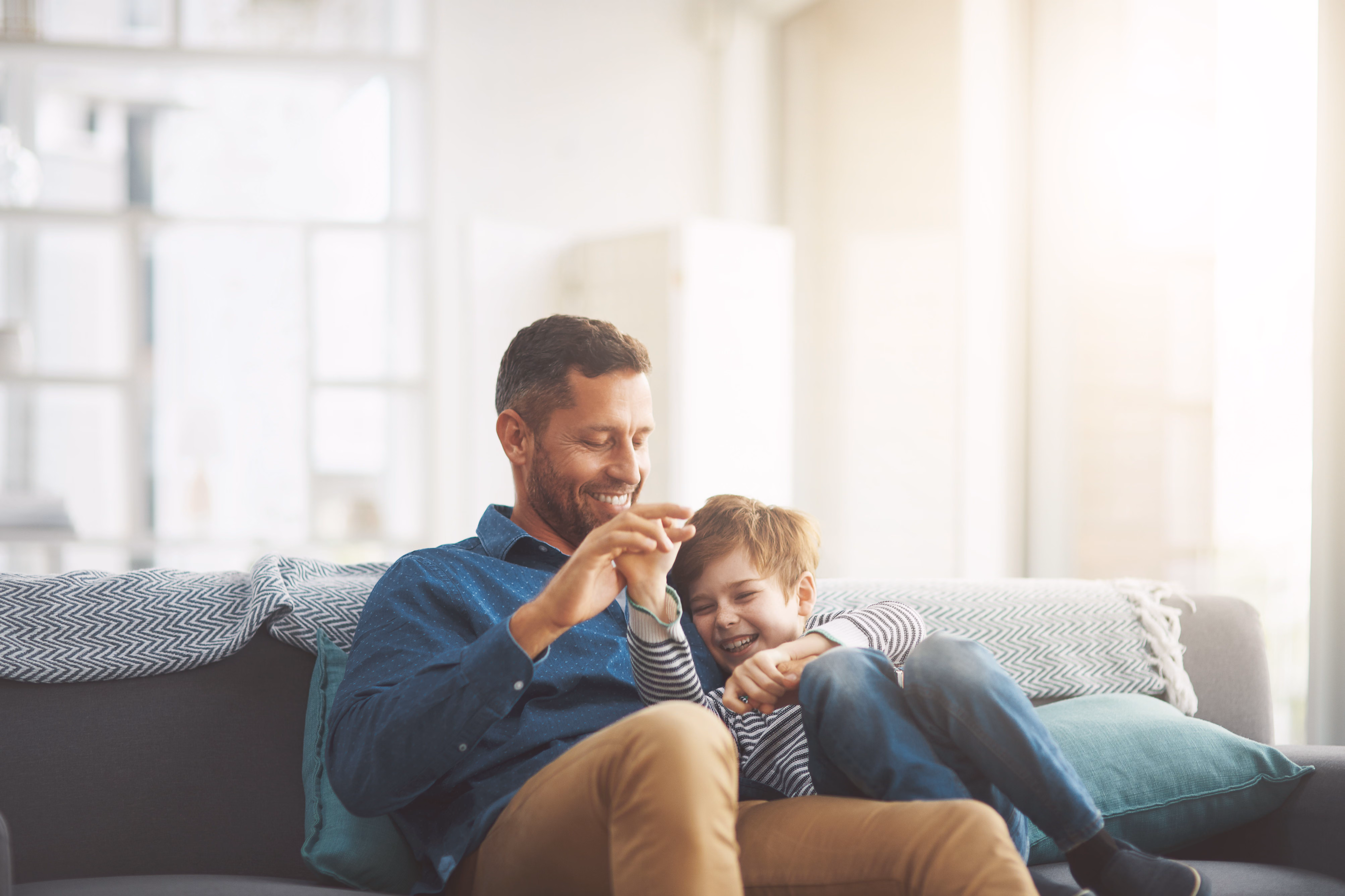 Smiling father playfully holding hands with laughing young son sitting on a couch.