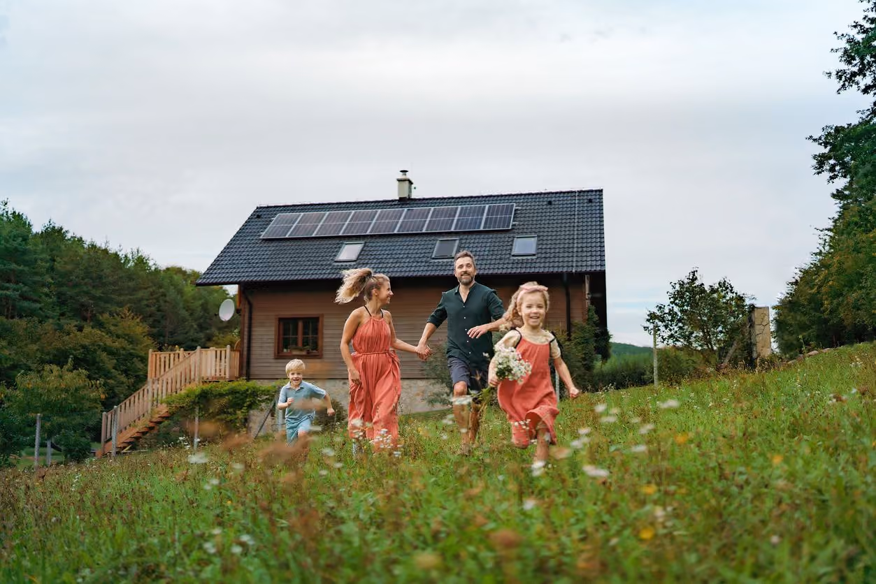 Family of four running and smiling in a wildflower meadow in front of a house with solar panels.