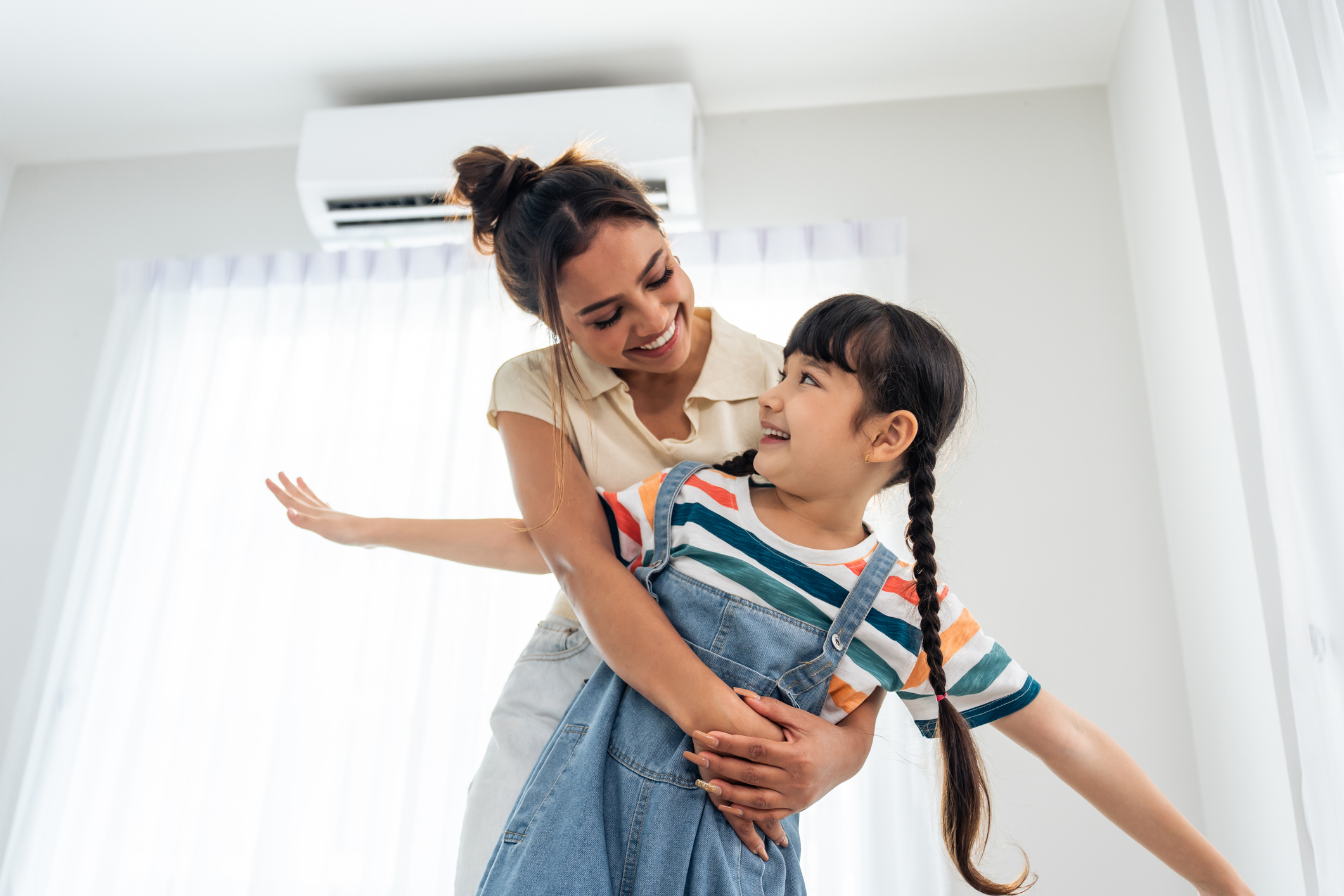 Smiling woman hugs and plays with a joyful girl with braided hair in a bright room.