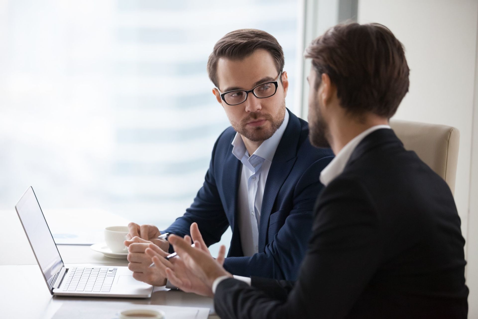 Two brokers in suits in a serious conversation at an office desk with a laptop and coffee cup