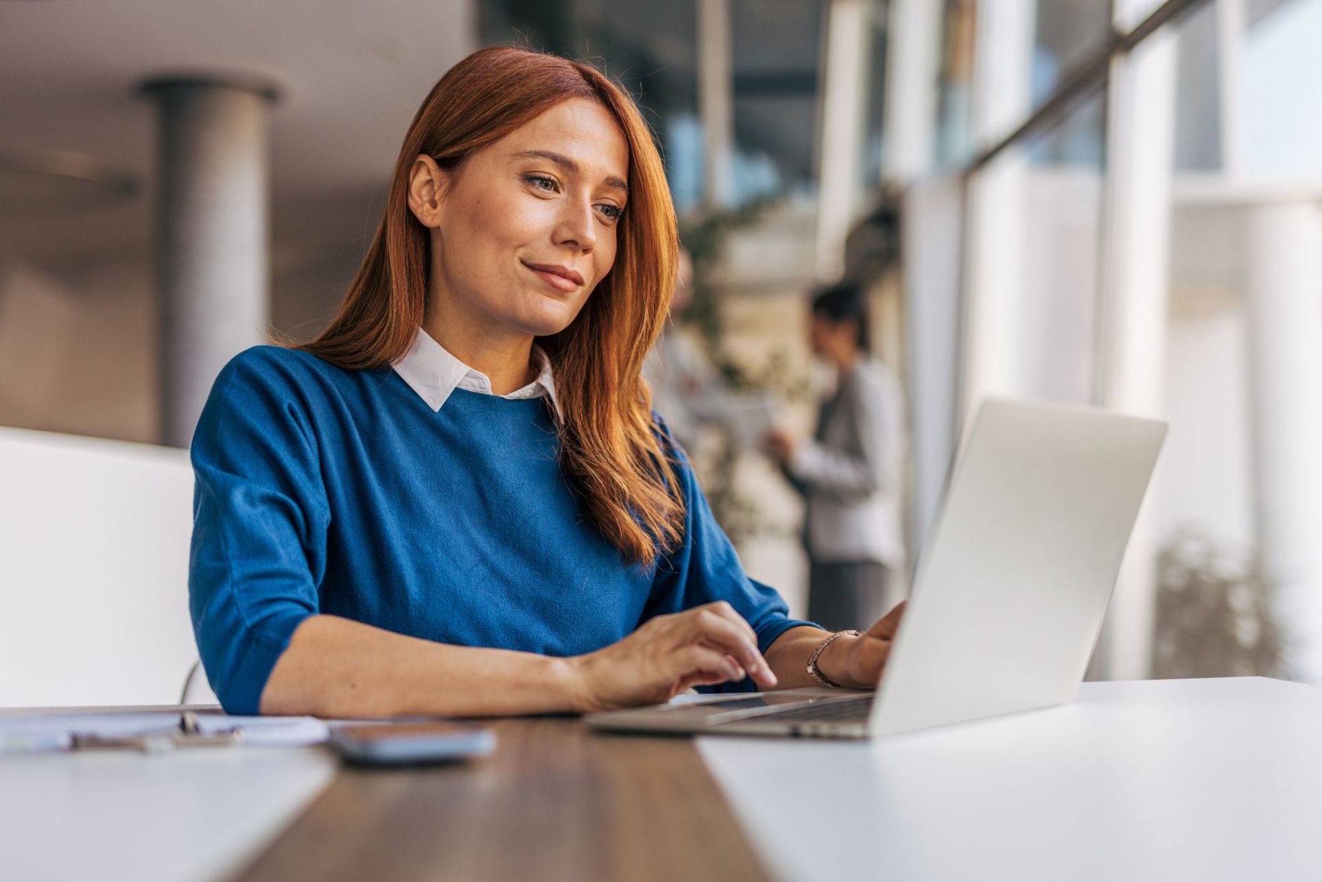 A woman with red hair sitting at a table typing on a laptop in a bright office setting