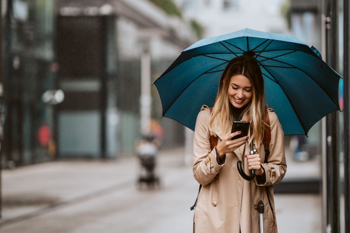 A woman walking outdoors under a blue umbrella in the rain while checking her phone
