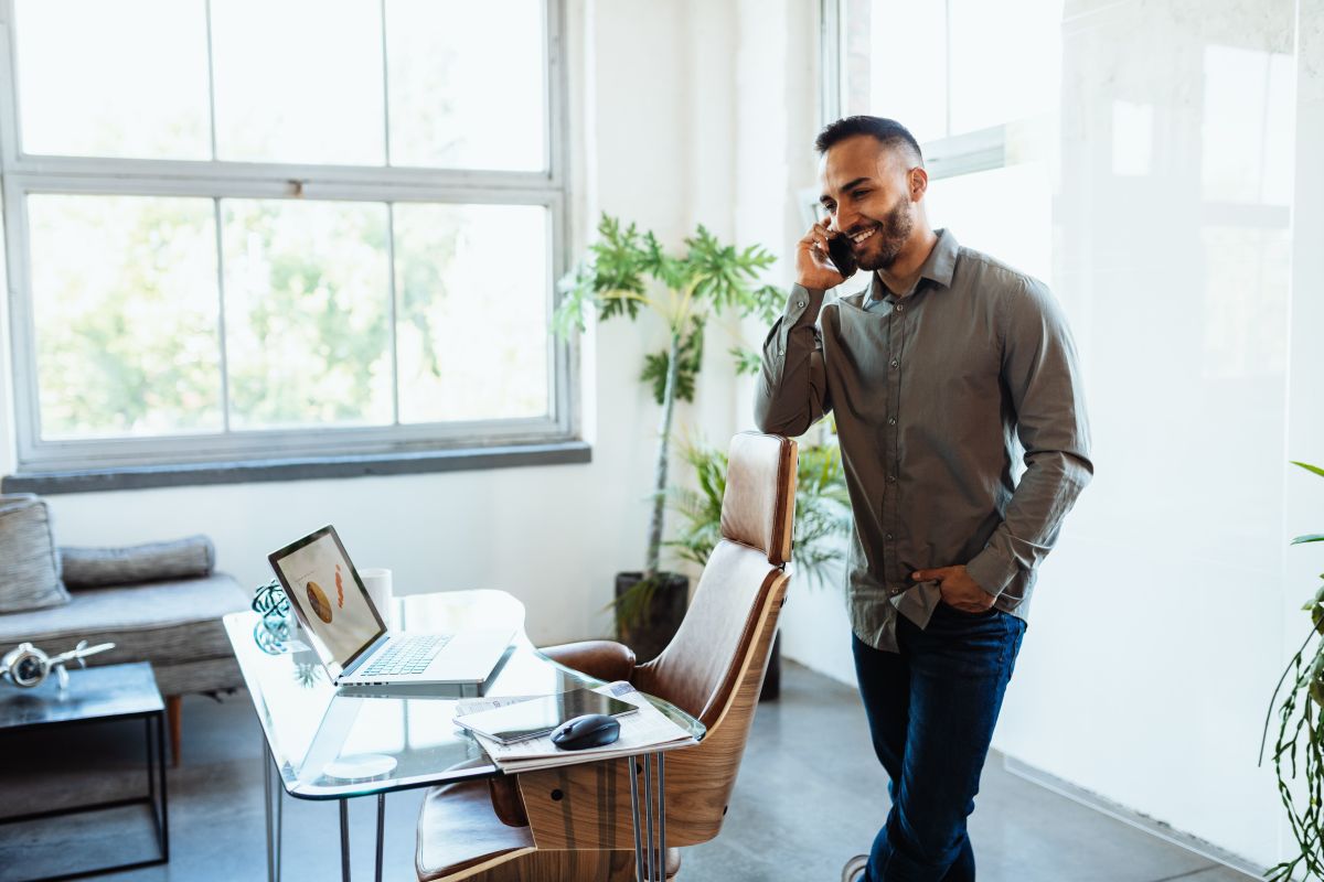 A man on a phone call standing beside a home office desk with a laptop and indoor plants