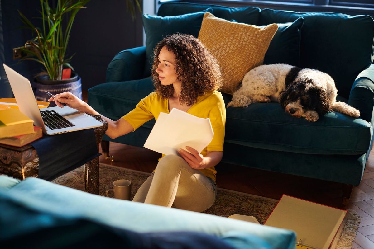 A woman sitting on the floor reviewing paperwork next to a laptop, with a dog resting on the couch behind her