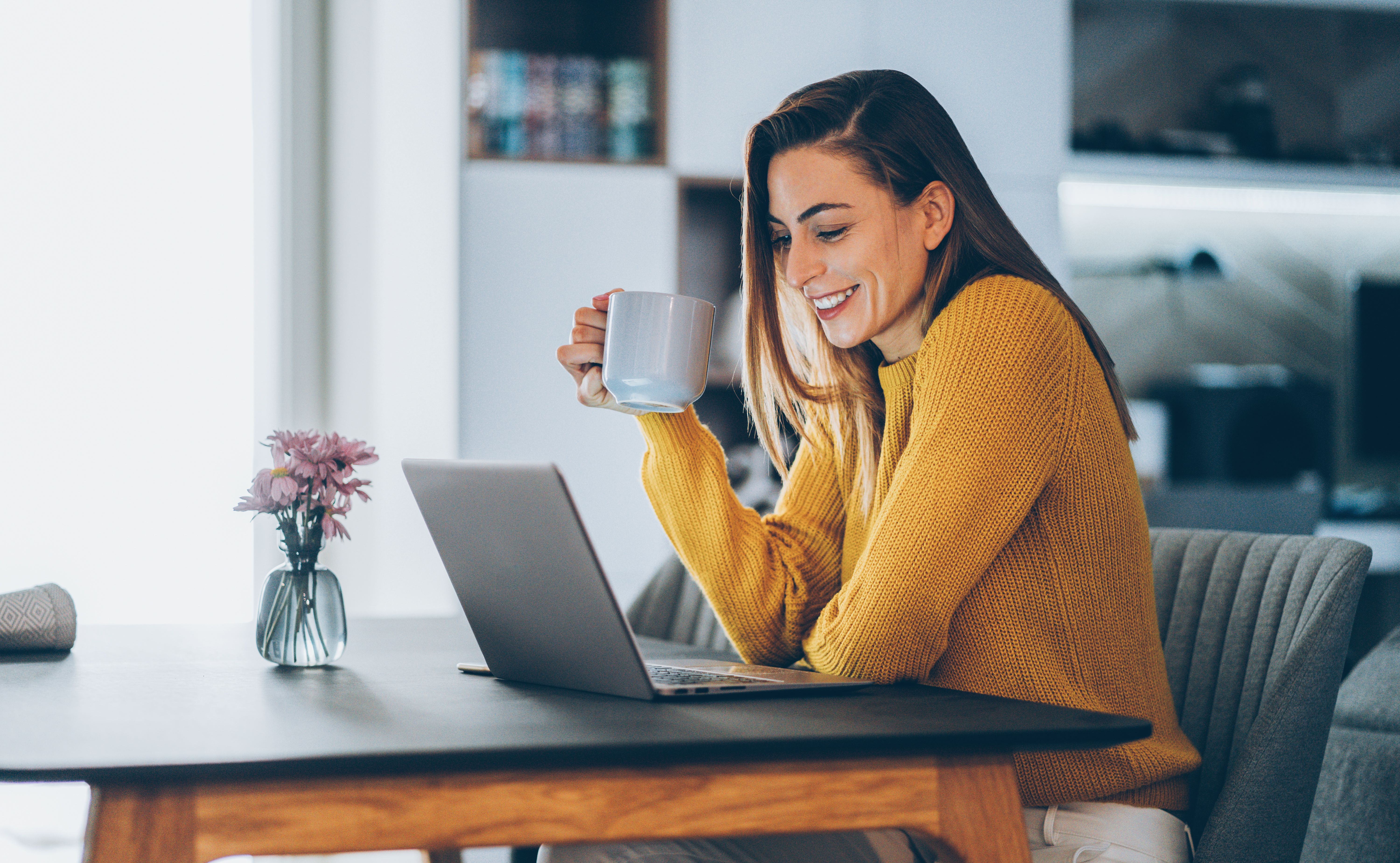 A woman in a yellow knit jumper sits at a table, smiling at her laptop while holding a mug