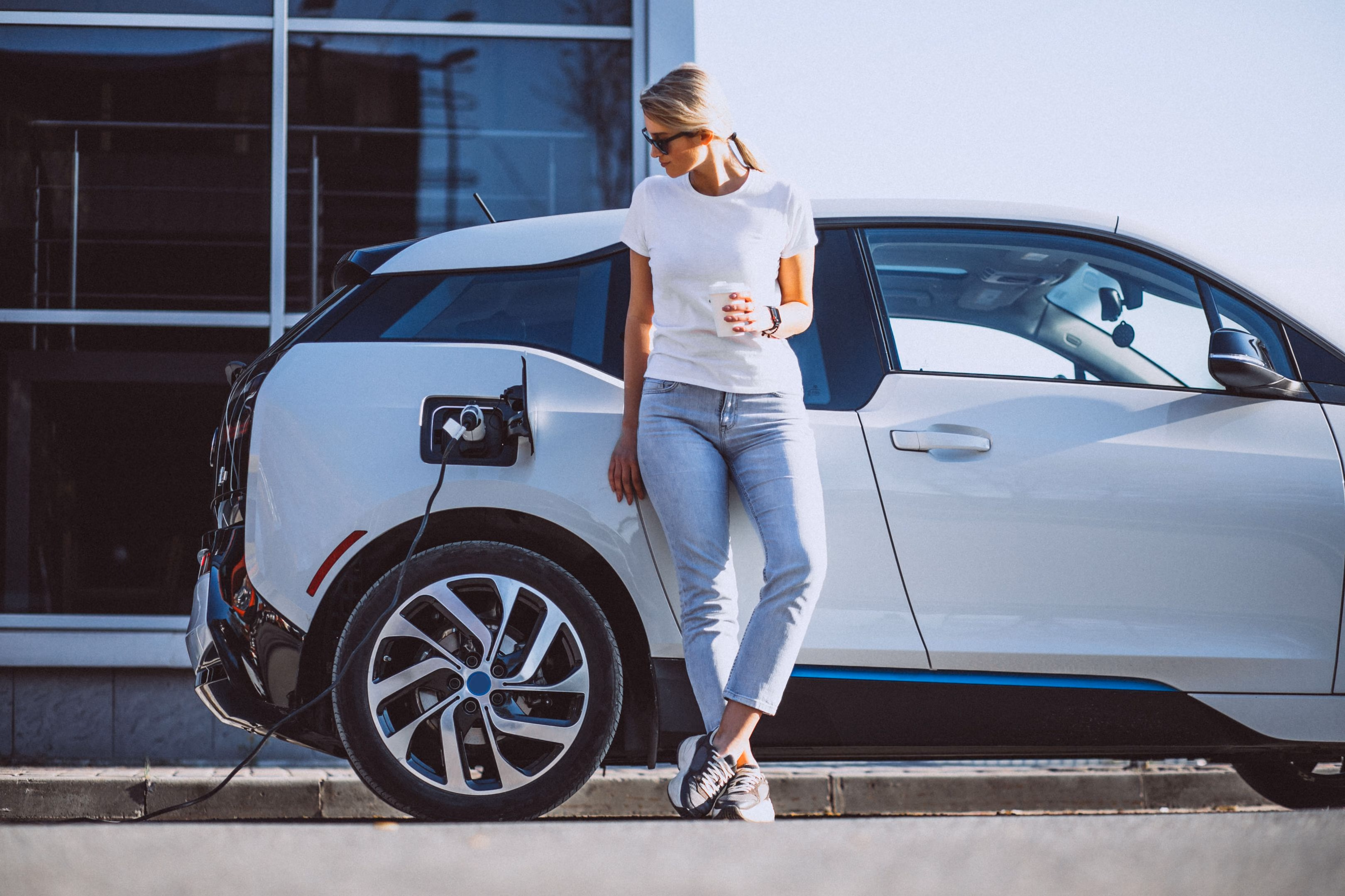 A woman in a white t-shirt and jeans leans against a white electric vehicle while it charges, holding a coffee cup