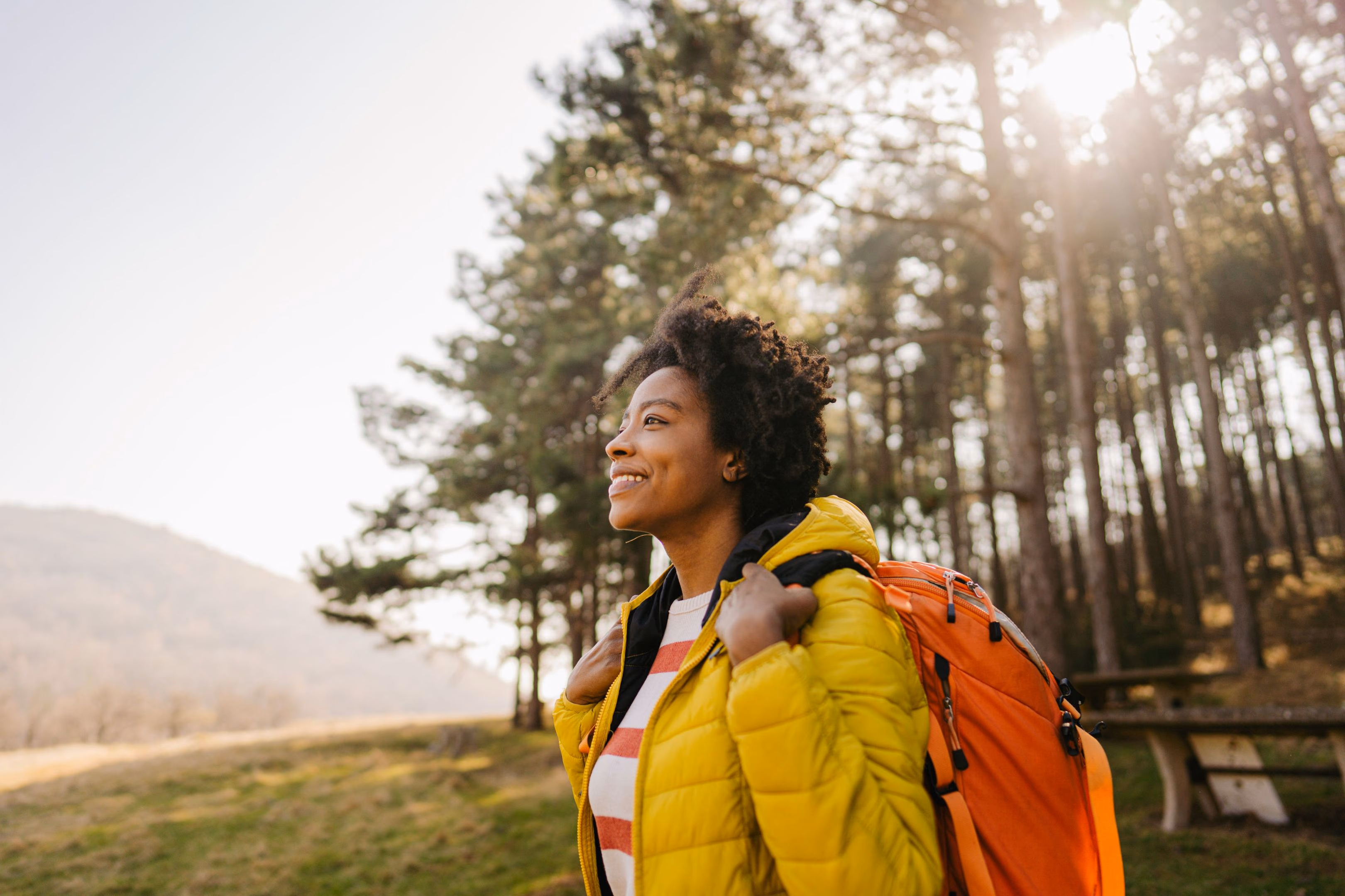 Smiling woman in yellow jacket with orange backpack hiking in a sunny forest