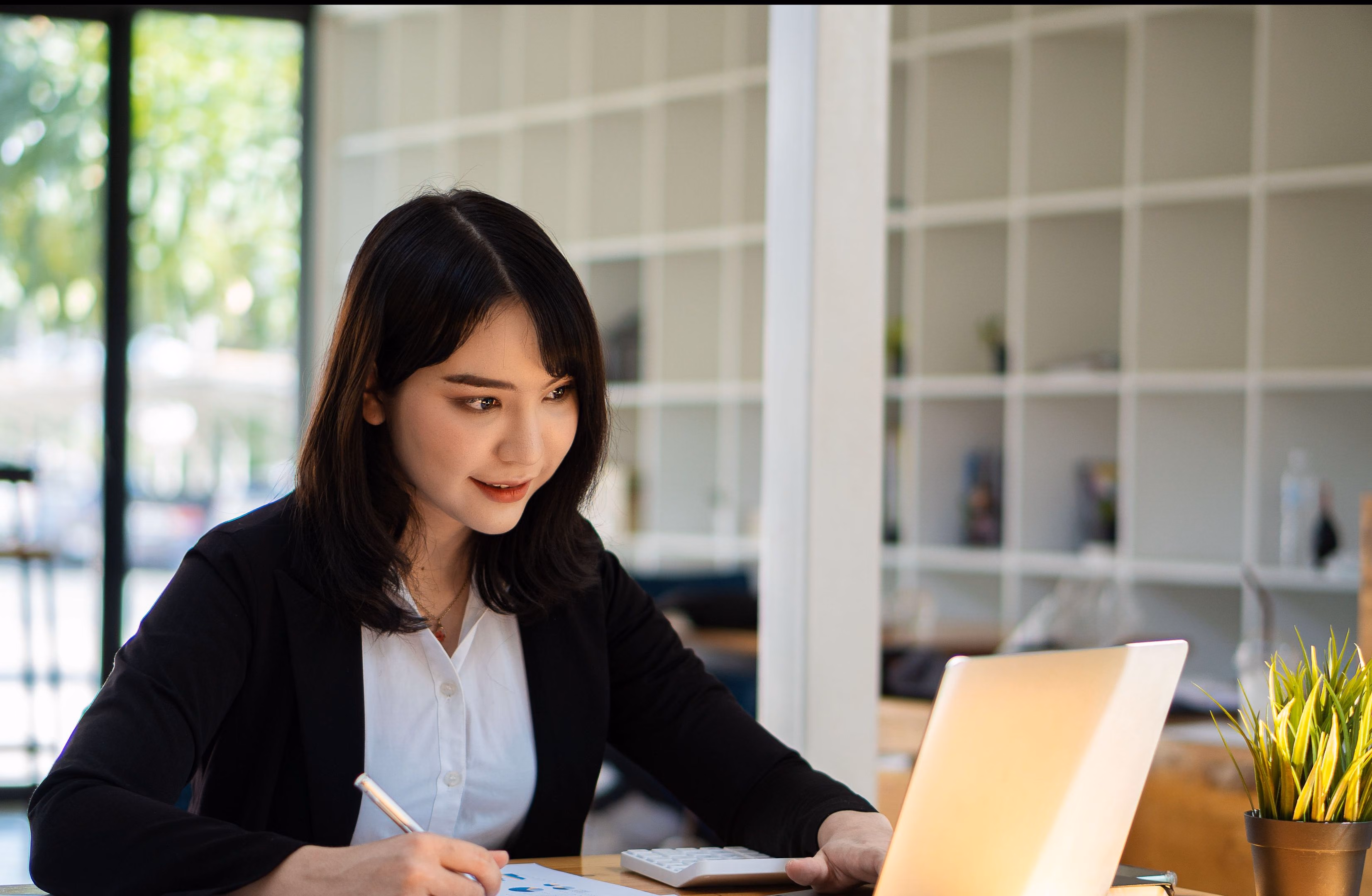 A woman in a black blazer sits at a desk using a calculator and pen while looking at a laptop