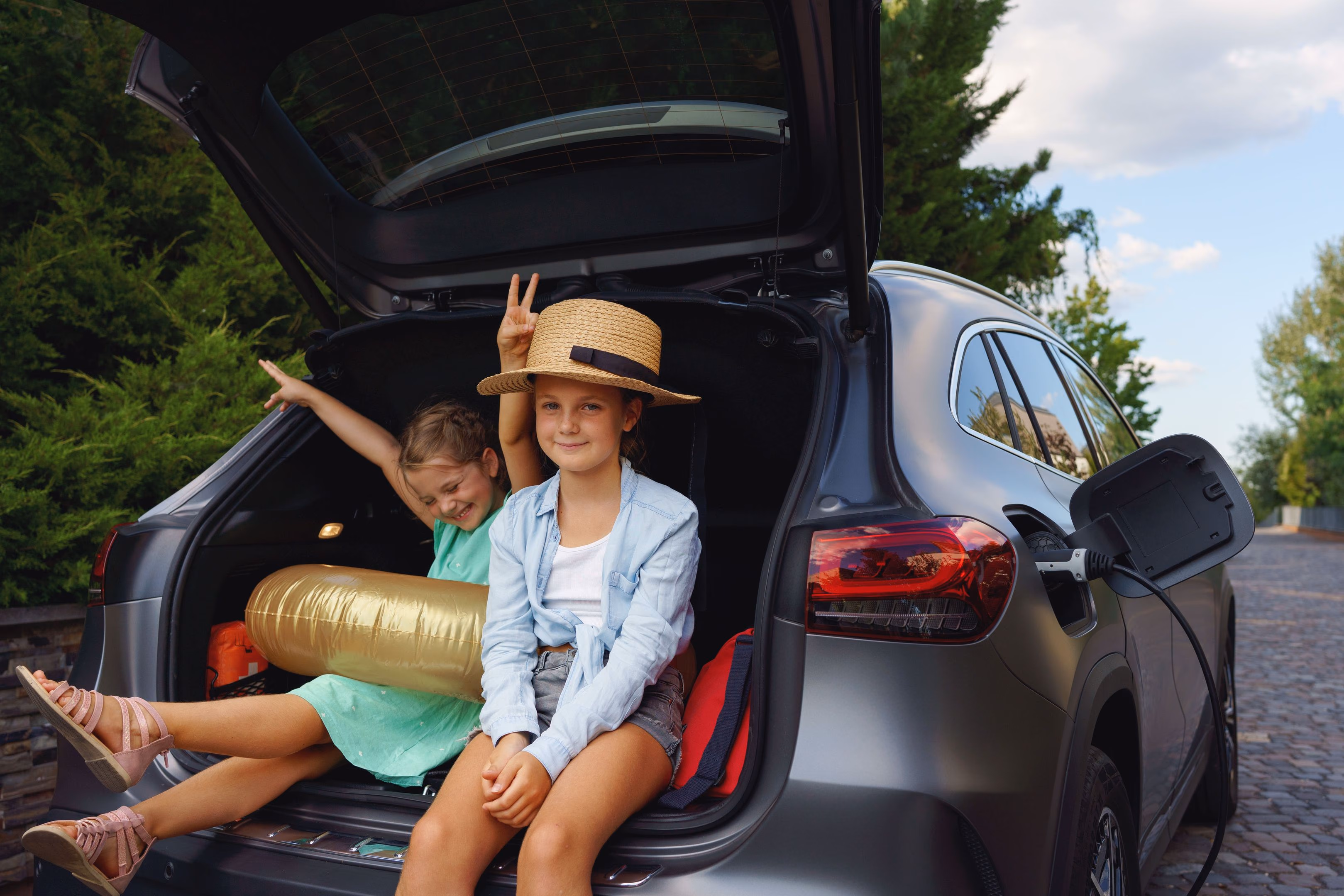 Two girls sitting in the open trunk of a grey electric car with its charging port plugged in