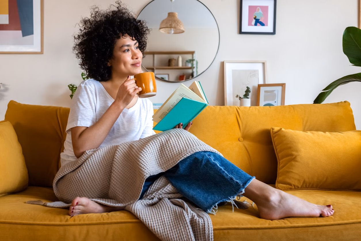 A person with curly hair sits on a yellow sofa, wrapped in a blanket, drinking from a mug while reading a book