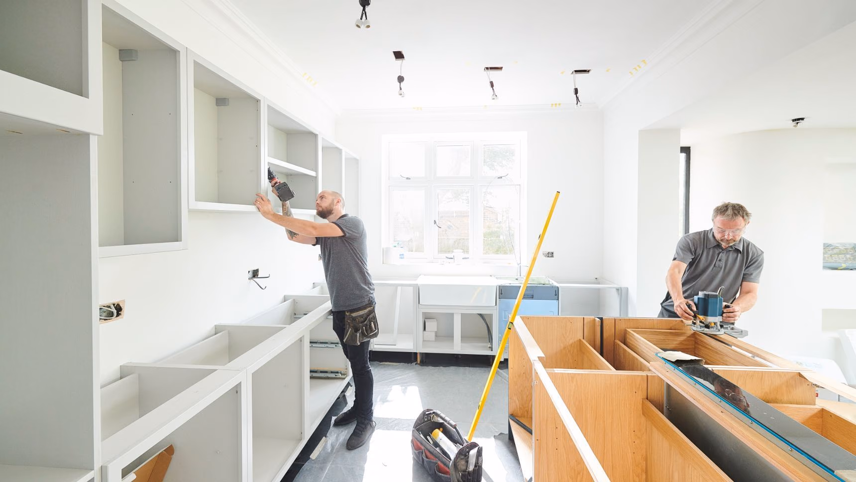 Two men fitting kitchen cabinets and worktops in a bright, unfinished white room